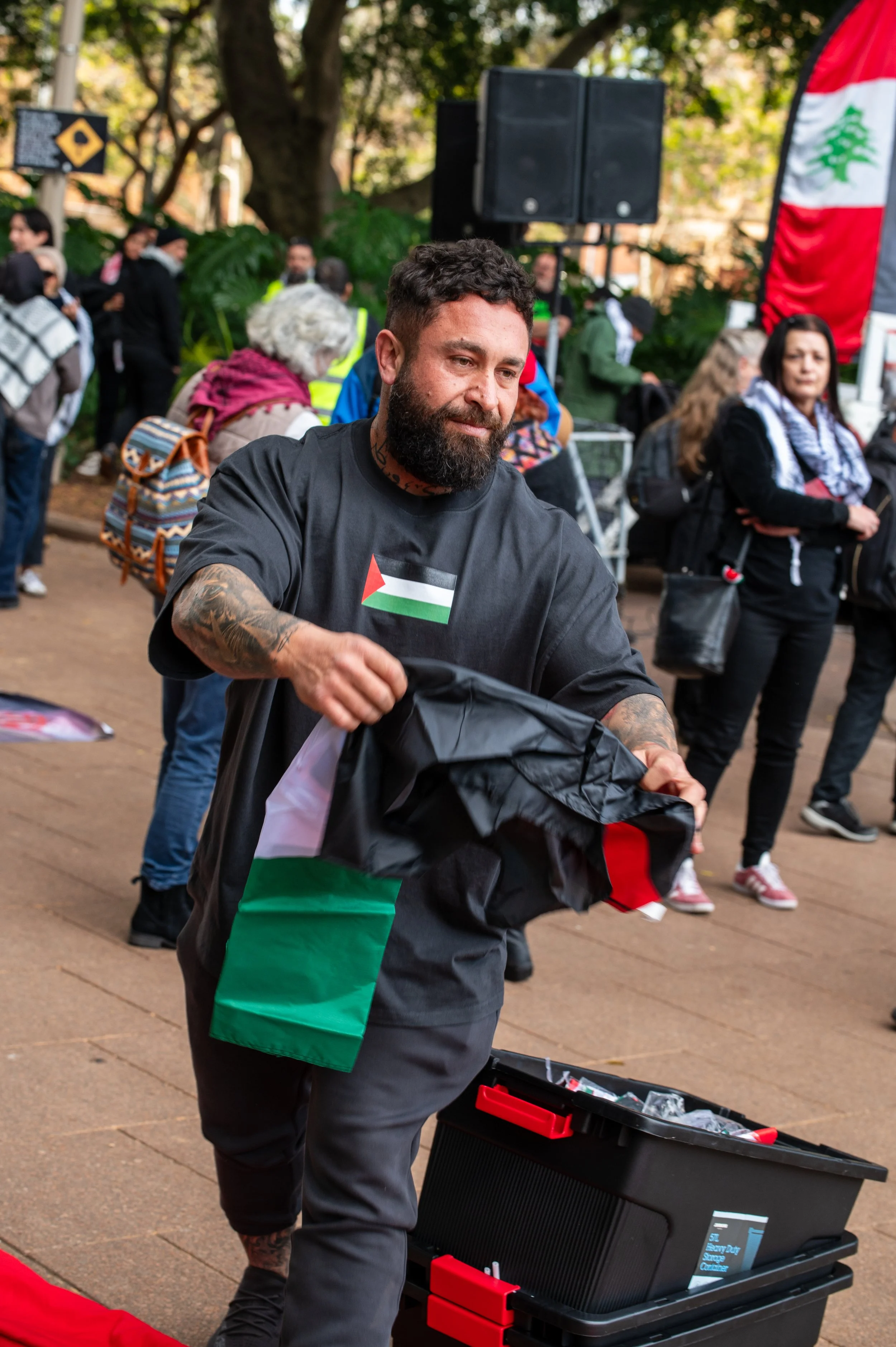 A man with a beard, tattoos, and wearing a black T-shirt with a Palestinian flag, holding a jacket with the same flag, at an outdoor event with multiple people, greenery, and speakers in the background.