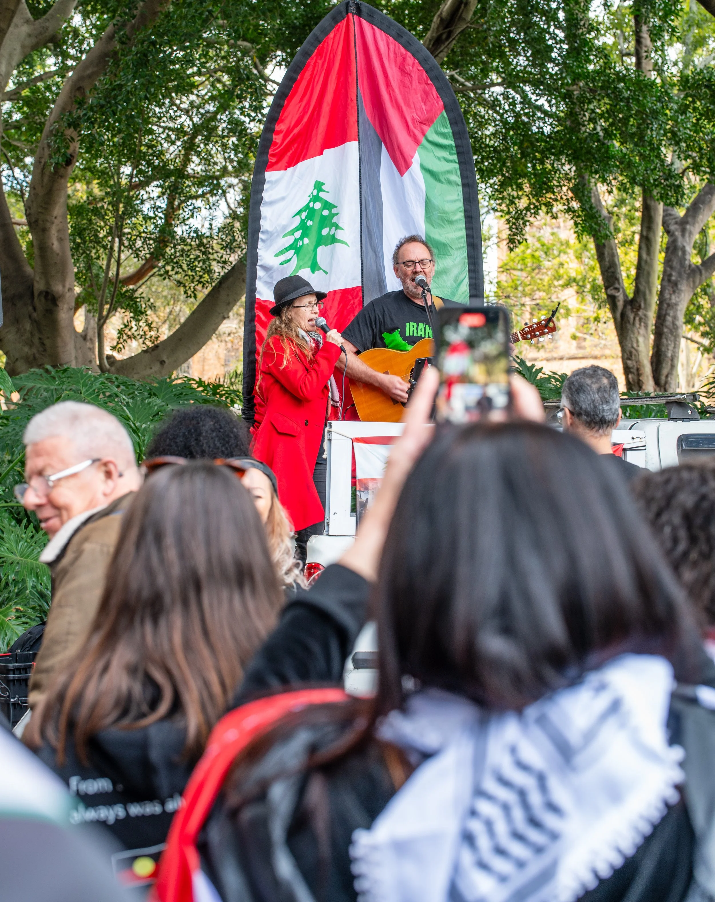 Two performers are singing and playing guitar on a stage with a Lebanese flag backdrop, during an outdoor event surrounded by an audience.