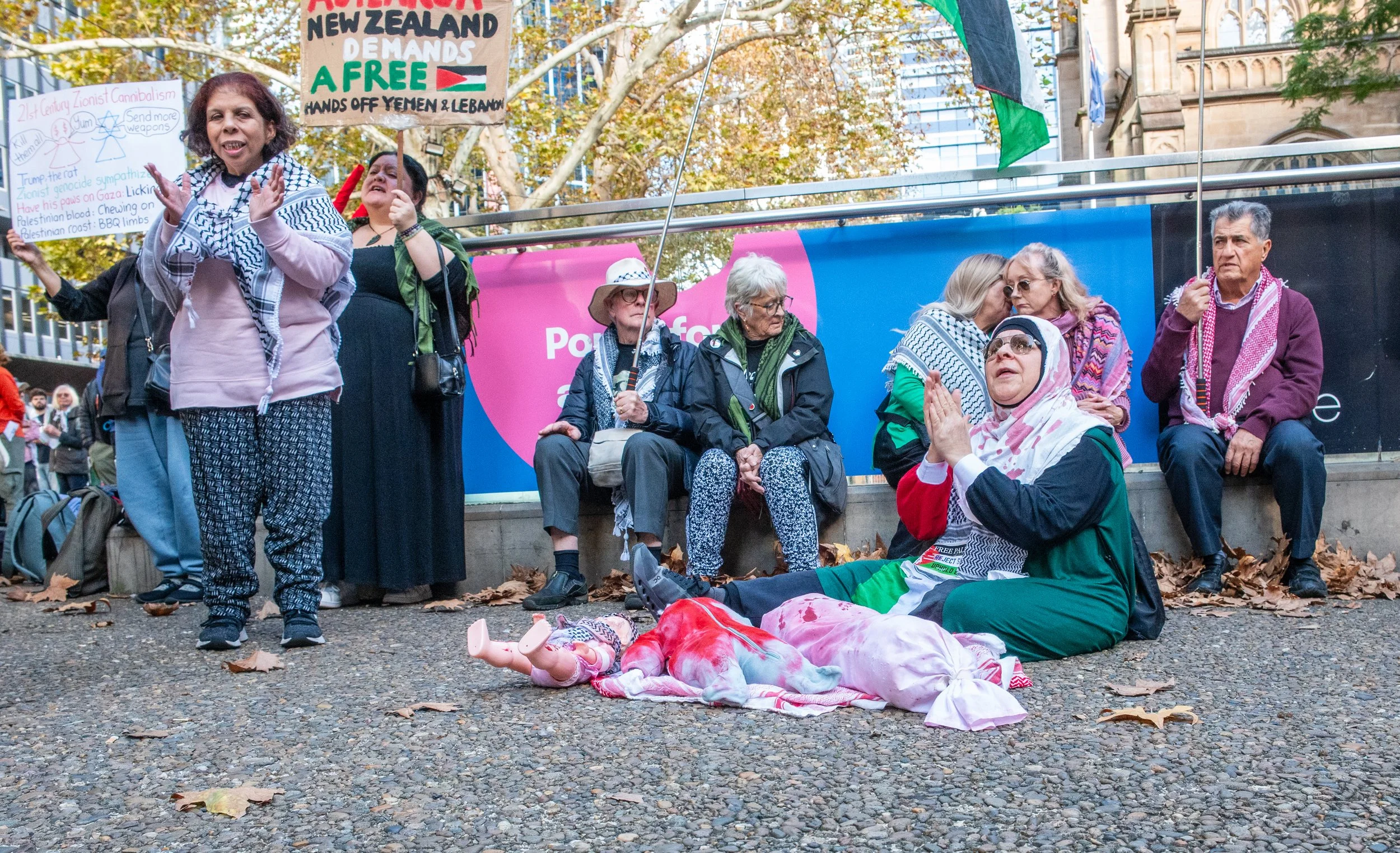 Group of protestors sitting and standing on a city street with signs and flags, participating in a demonstration for Palestine and against violence in Yemen and Lebanon. One woman kneels on the ground with hands clasped, wearing a headscarf and glasses. The ground is scattered with fallen leaves.