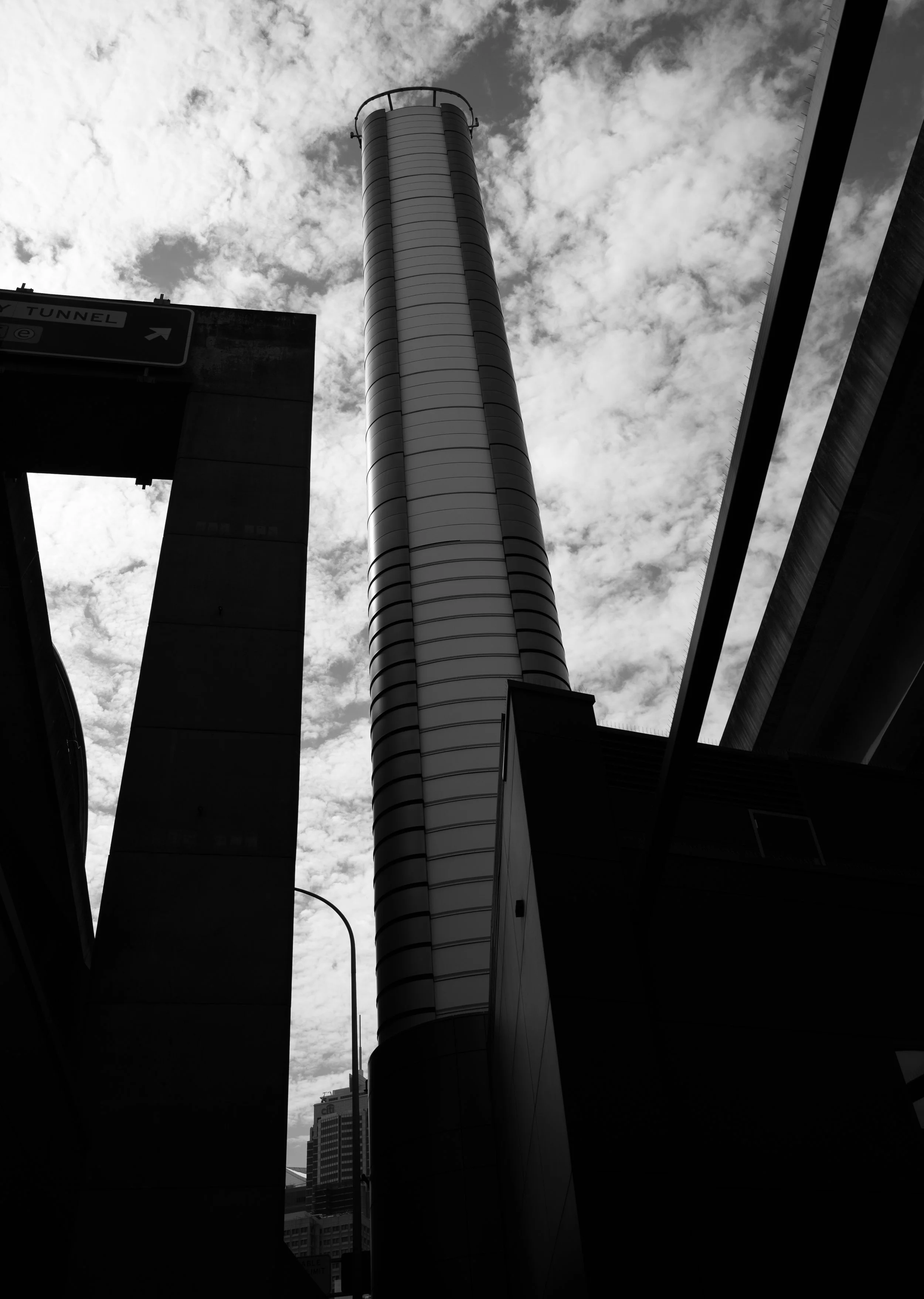 A black and white photo of a tall, cylindrical skyscraper with a circular platform at the top, partially obscured by surrounding buildings and an overcast sky with scattered clouds.