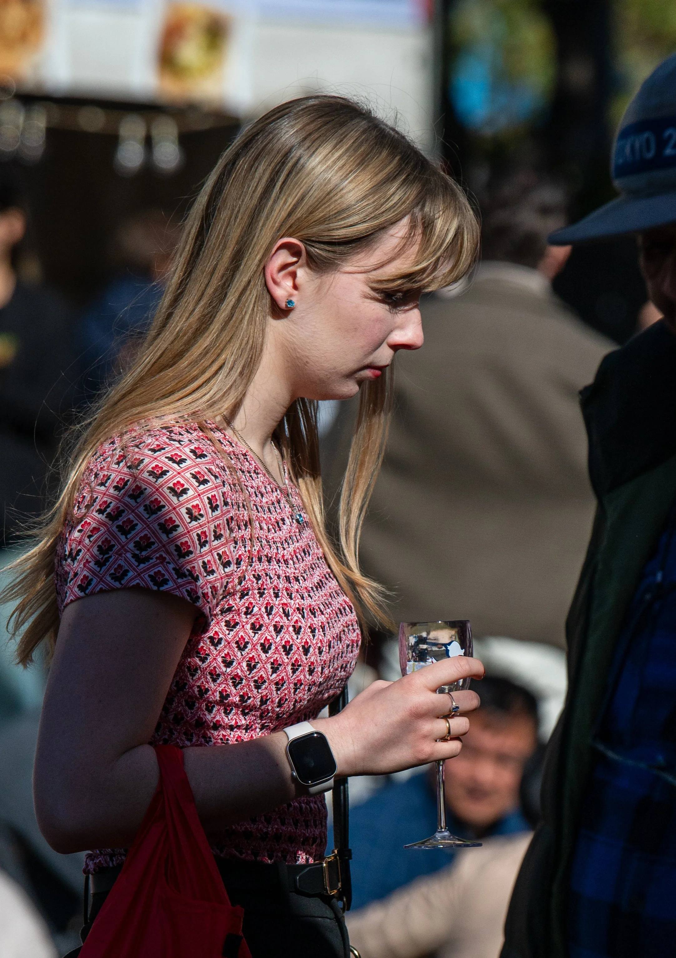 A young woman with long blonde hair wearing a patterned pink and red top, a smartwatch, and earrings, holding a glass of wine at an outdoor event.