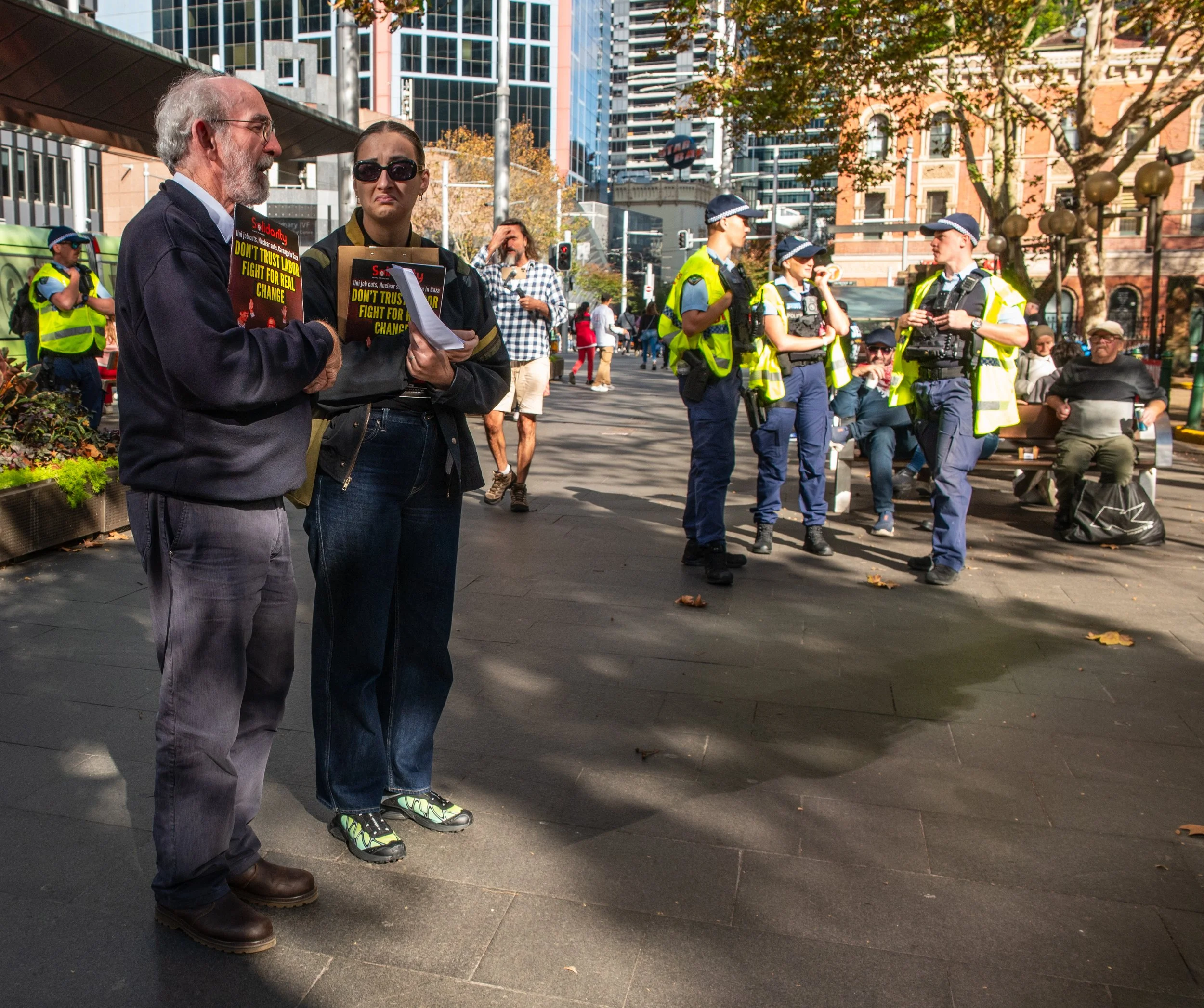 Two people holding signs and brochures speaking in front of police officers and seated individuals in a city square with tall buildings and trees.