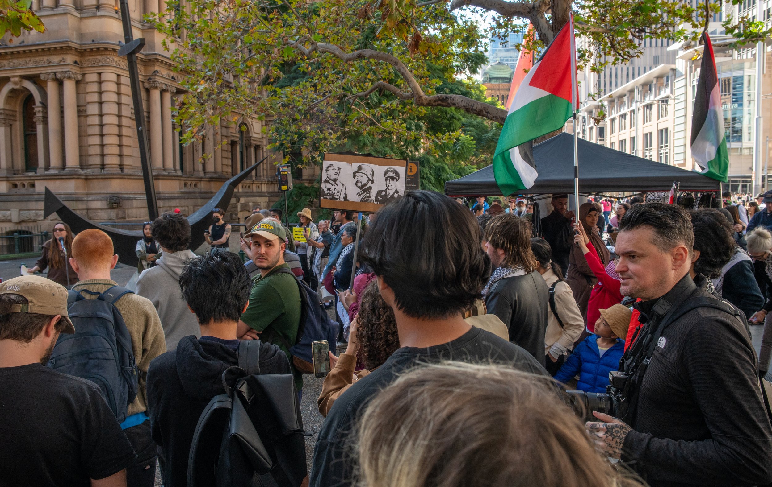 Crowd of protesters gathered outdoors, holding Palestinian flags and a photograph of historic figures, with city buildings and trees in the background.
