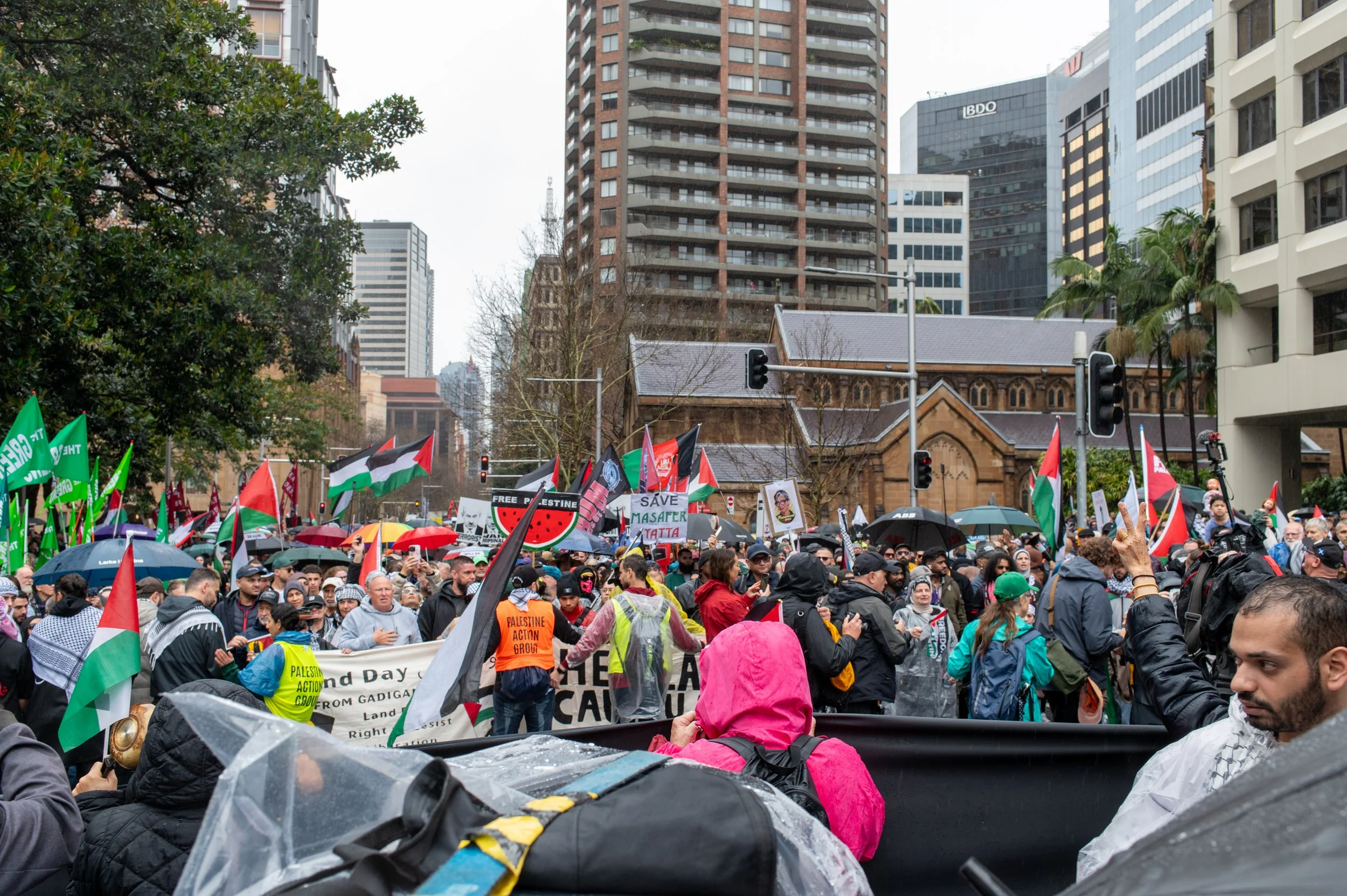 Crowd of protestors holding flags and signs in an urban setting with high-rise buildings, some people holding umbrellas, and a historic church-like building in the background during daytime.
