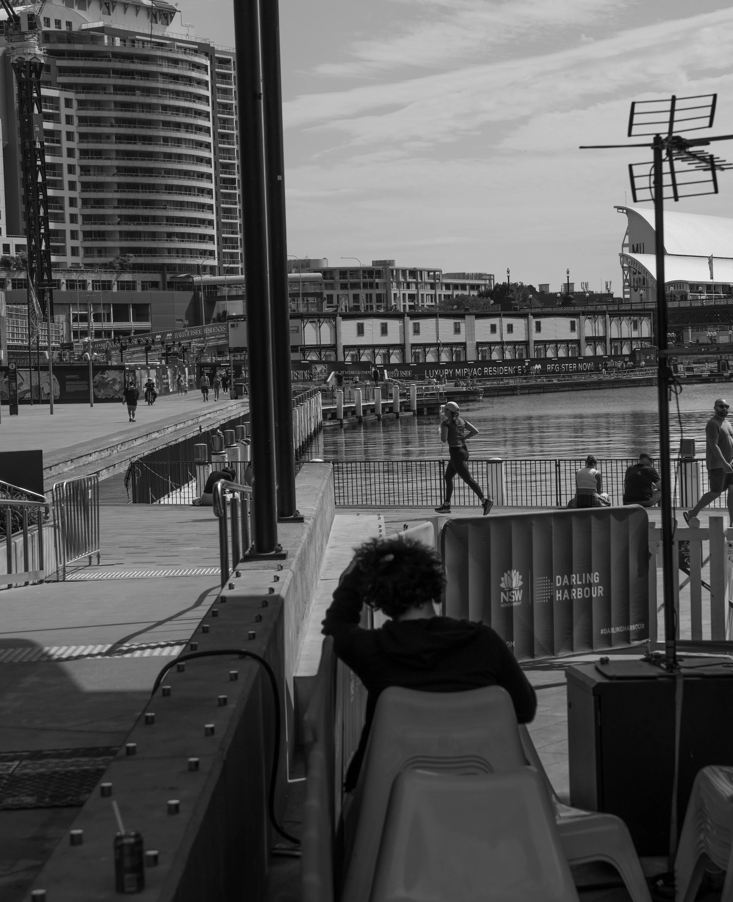 A black and white photo of Darling Harbour in Sydney, featuring a waterfront promenade, modern high-rise buildings, and people walking or sitting by the water.