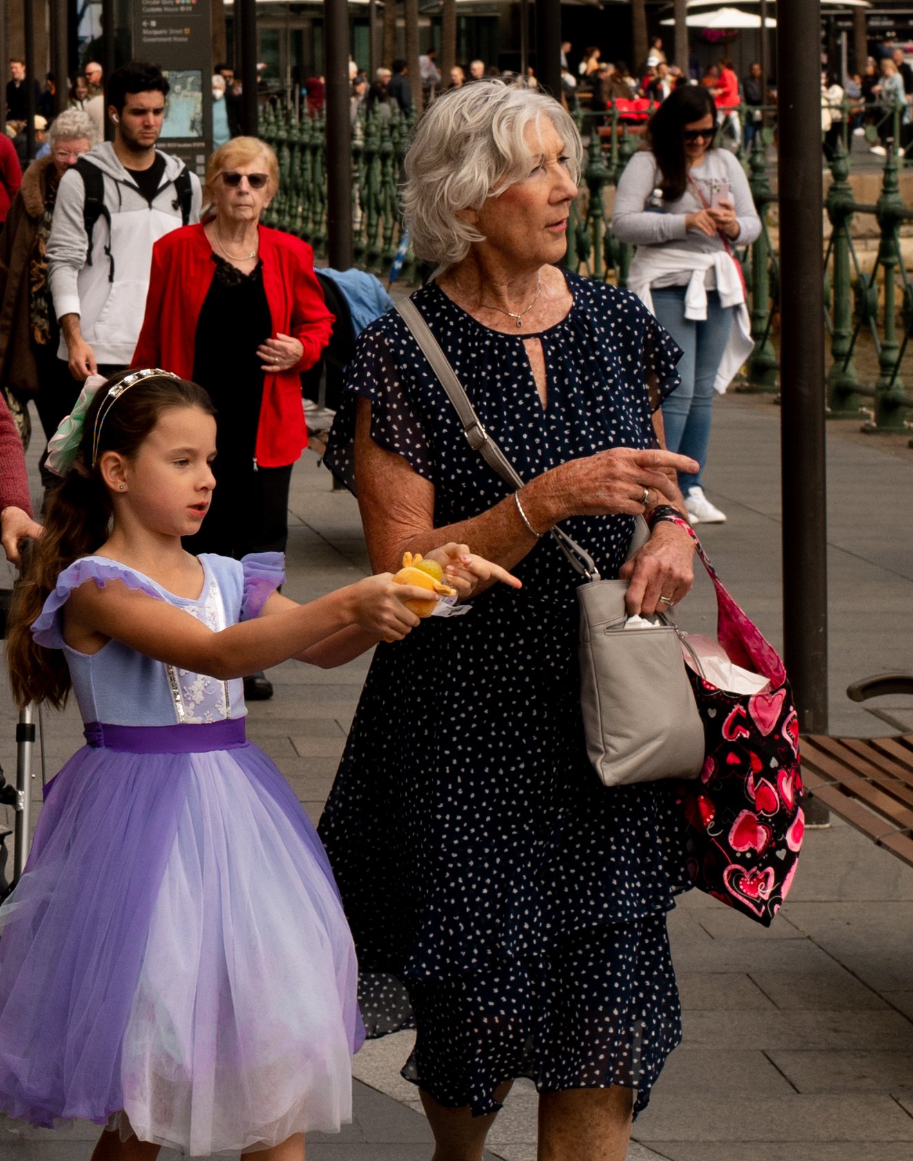 A young girl in a purple dress holding a yellow object, walking with an elderly woman in a black polka-dot dress in a crowded outdoor area.