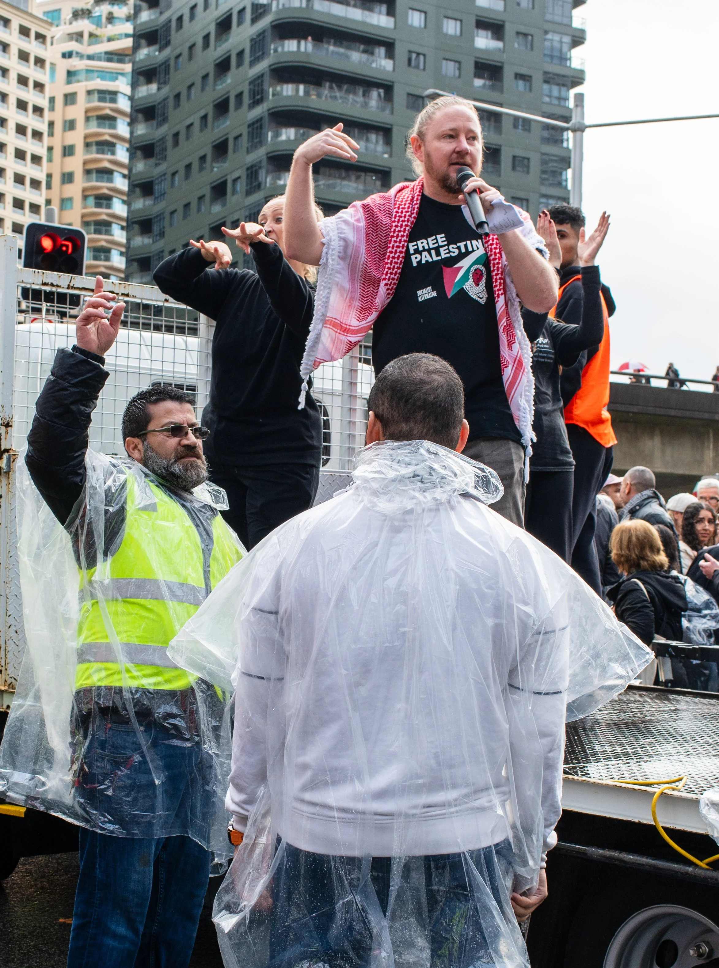 A man holding a microphone and speaking at a protest, wearing a "Free Palestine" shirt and a red-and-white scarf. He stands on a platform, surrounded by others, some wearing rain ponchos, with a cityscape of tall buildings in the background.