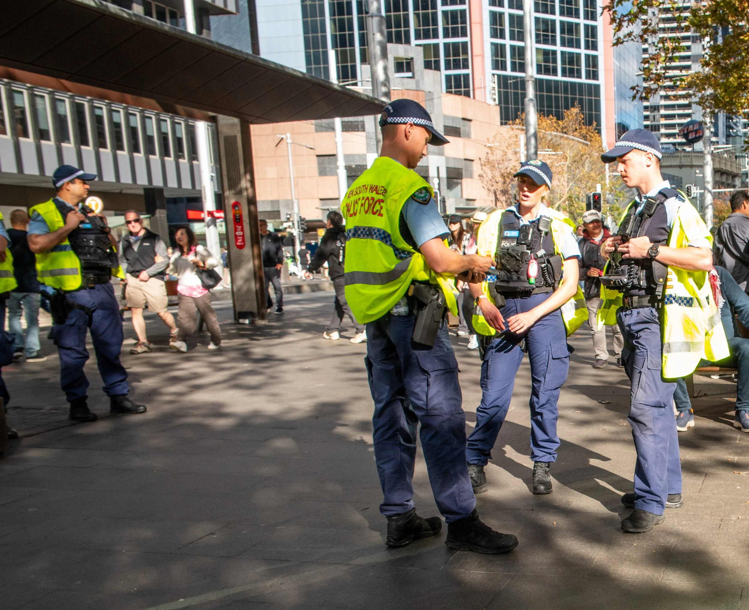 Police officers in yellow vests and blue uniforms standing and talking in a city street, with pedestrians walking in the background.