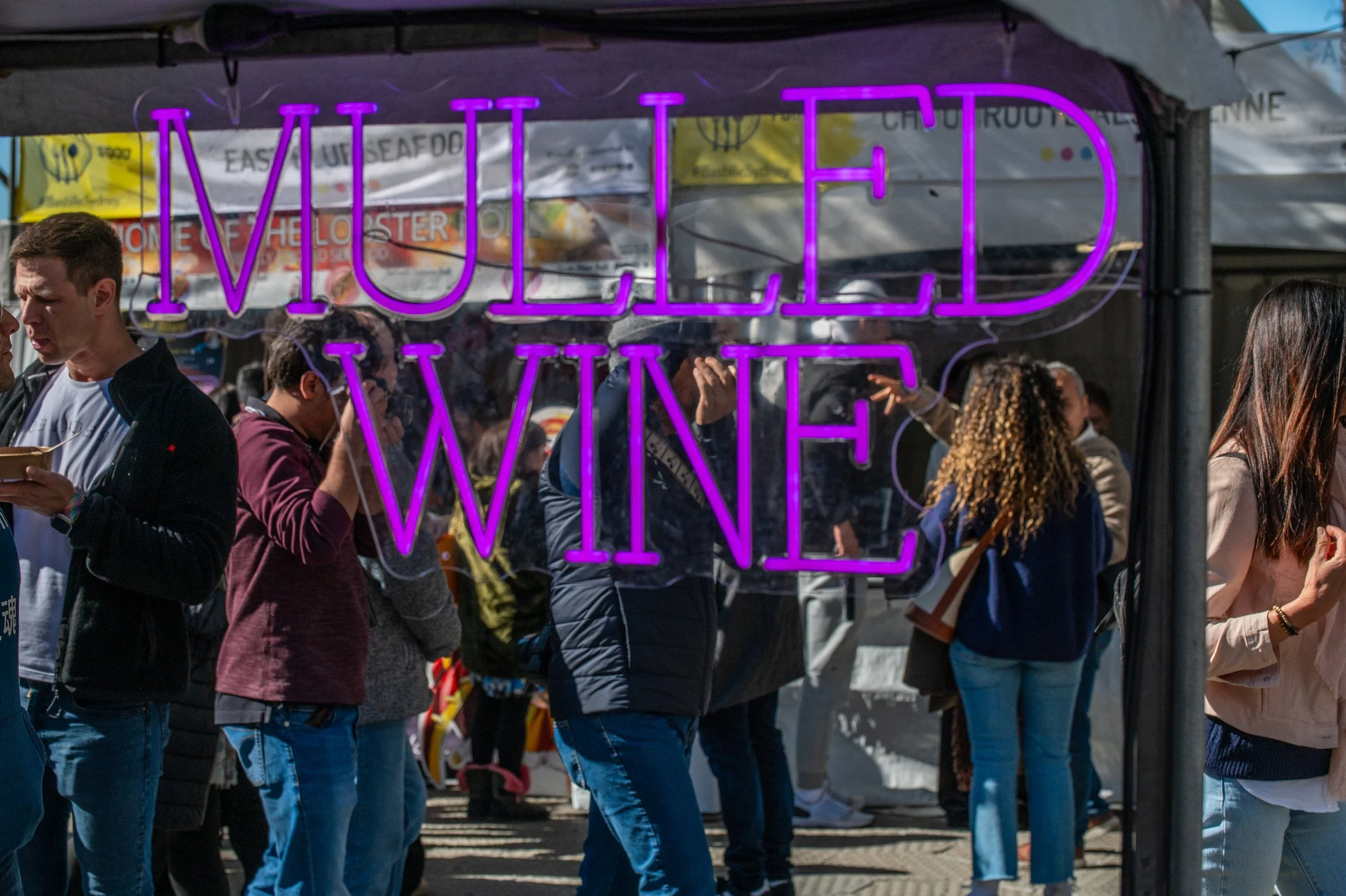 People gather at an outdoor event with a neon sign reading 'MELLED WINE' in purple above. Some are eating or talking, and there are food stalls and colorful banners in the background.