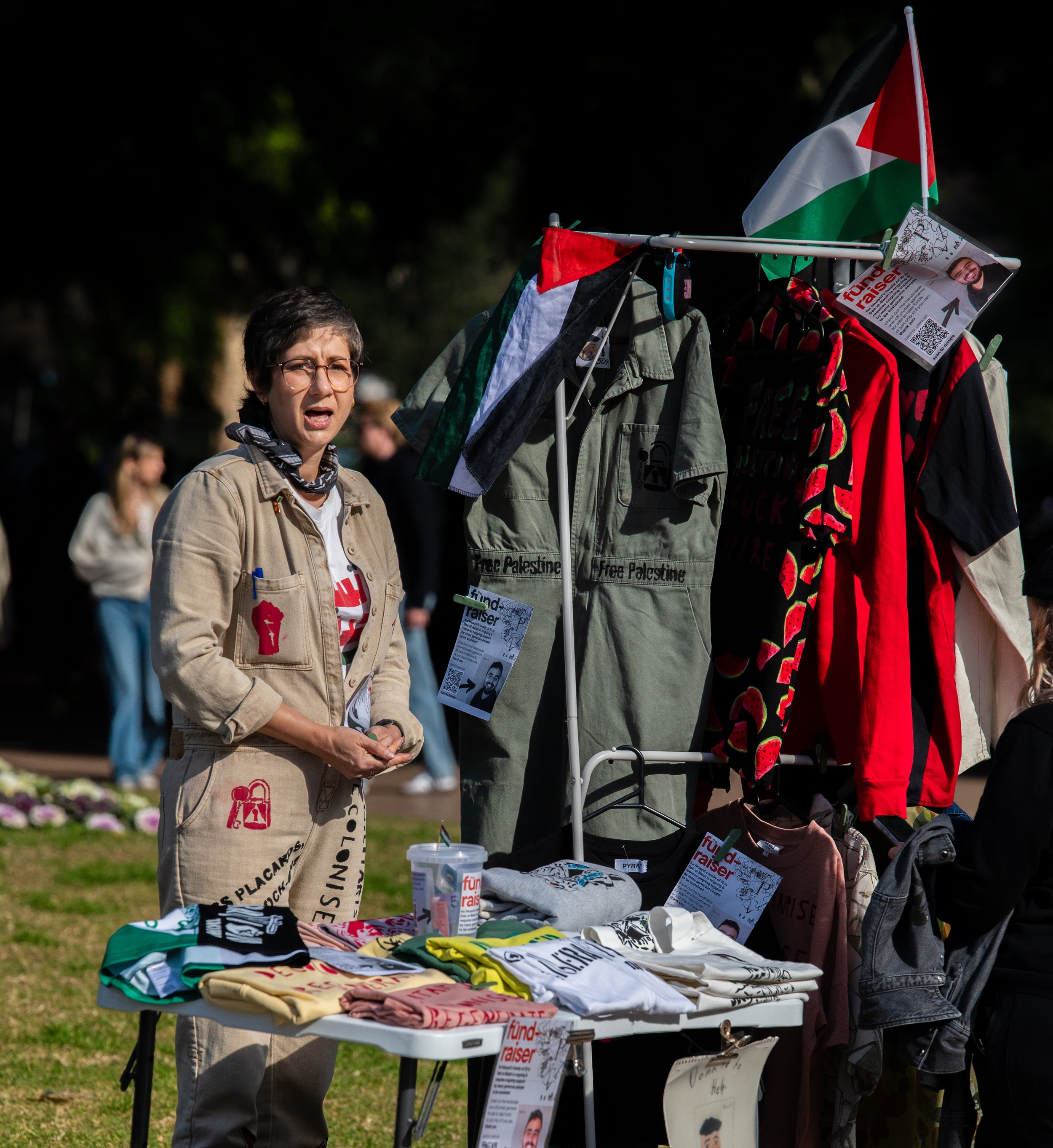 A woman at a protest booth with Palestinian flags, t-shirts, and signs, speaking to a crowd.
