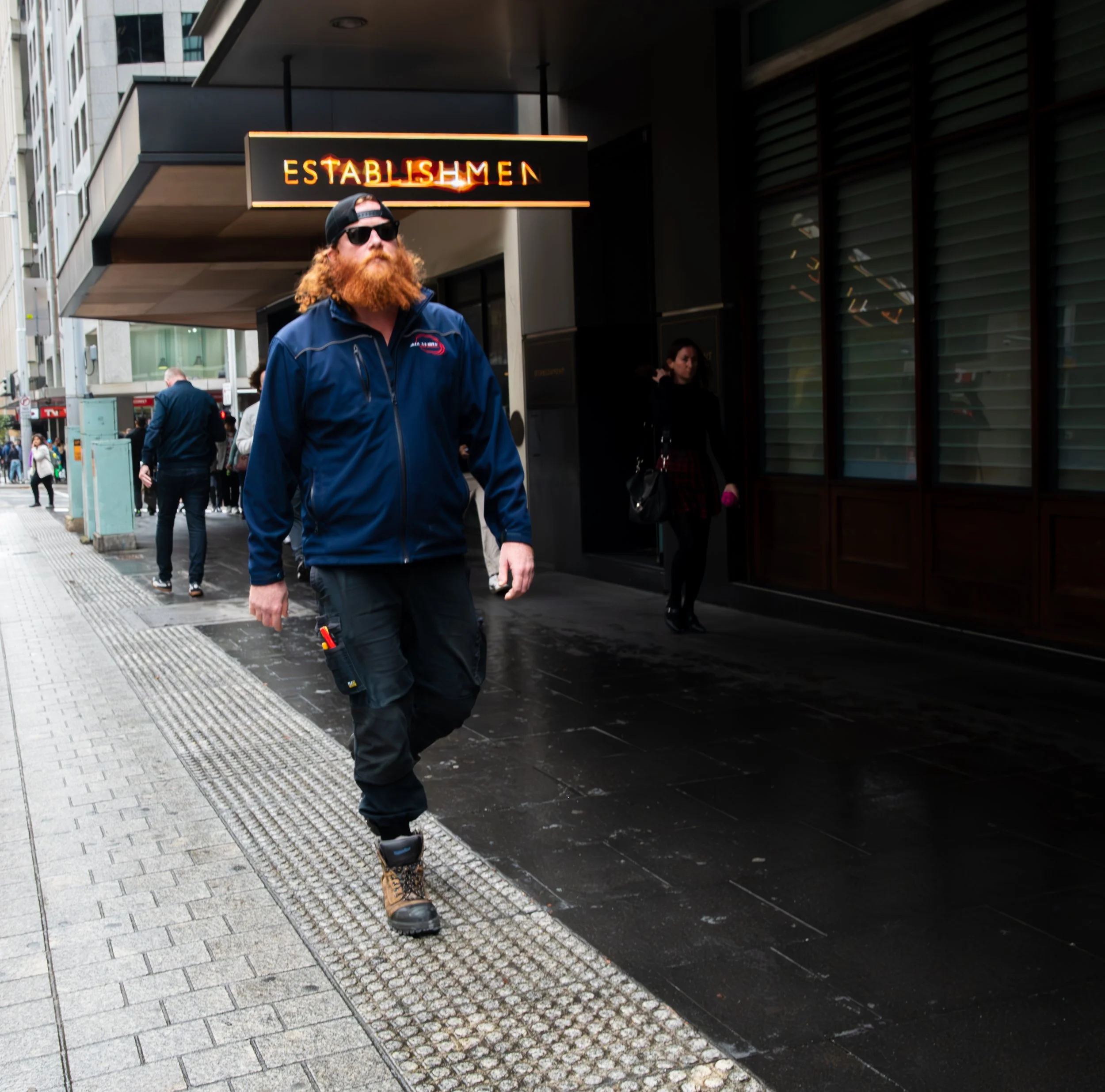 Man with red beard, sunglasses, and dark blue jacket walking on a city sidewalk near an establishment sign.