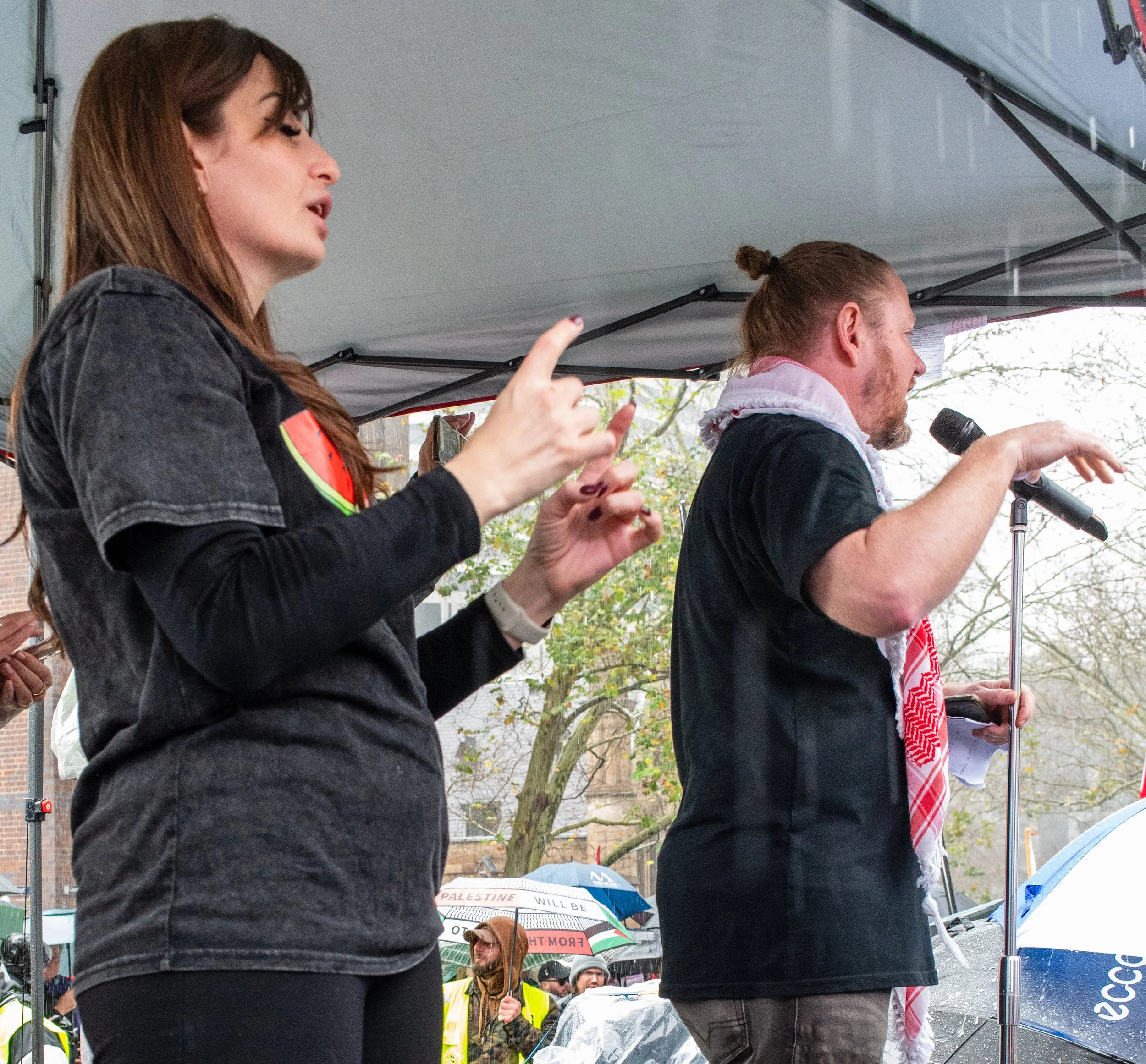 A woman and a man are speaking at a rally or protest under a tent. The woman is making a gesture with her hands, and the man is holding a microphone and reading from a paper. In the background, people are holding umbrellas and some are wearing yellow vests.