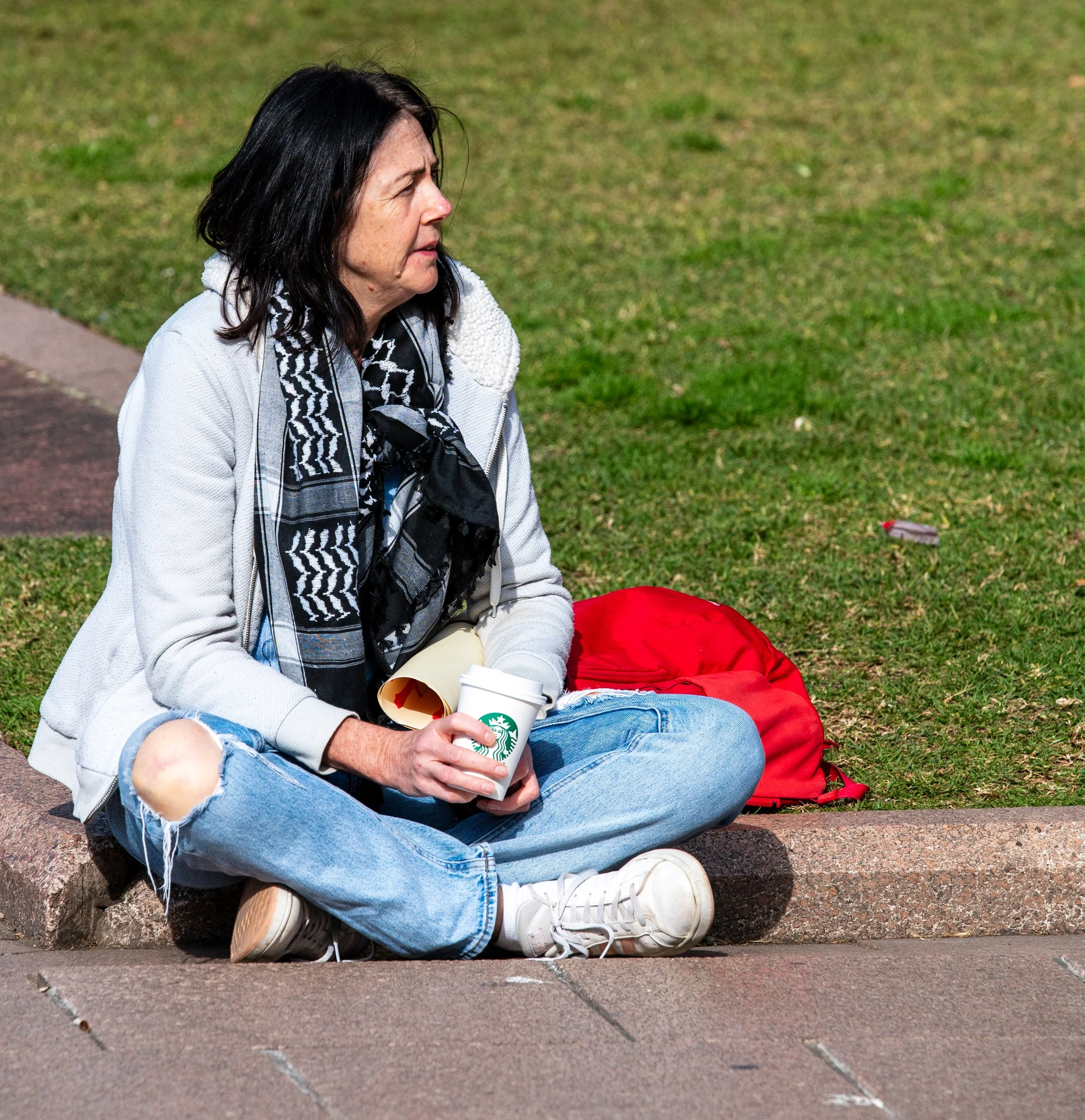 A woman sitting cross-legged on a curb, holding a Starbucks coffee cup, with a red backpack beside her on the grass, wearing a white jacket, distressed jeans, and a black-and-white patterned scarf.
