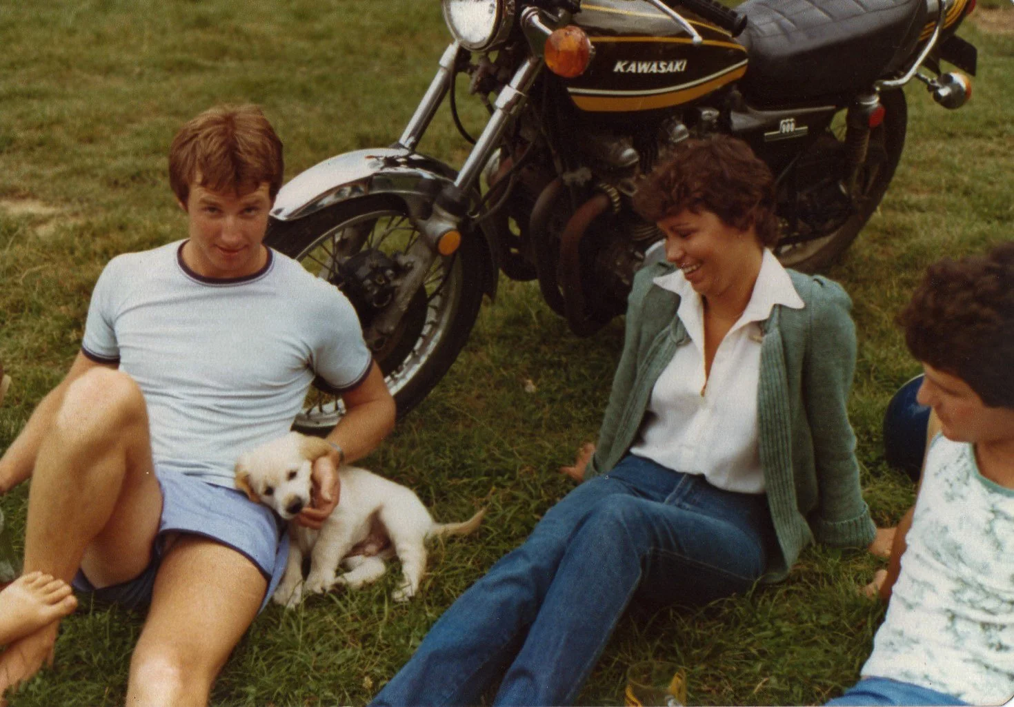 A group of people sitting on the grass, a man holding a dog, with a motorcycle in the background.