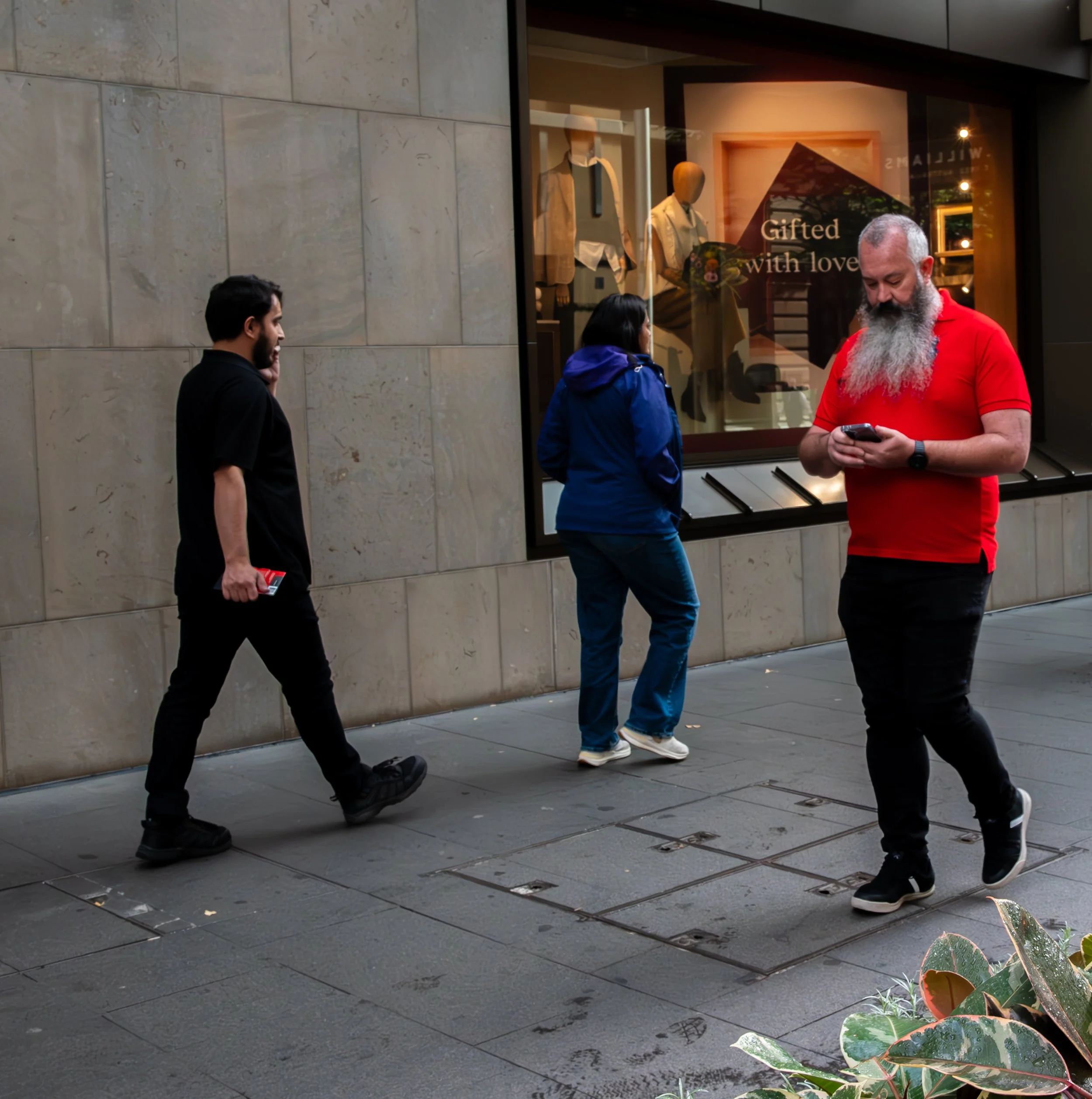 Three people walking on a city sidewalk with one person looking at their phone, two others walking past, in front of a storefront window that displays mannequins and a sign reading "Gifted with love."