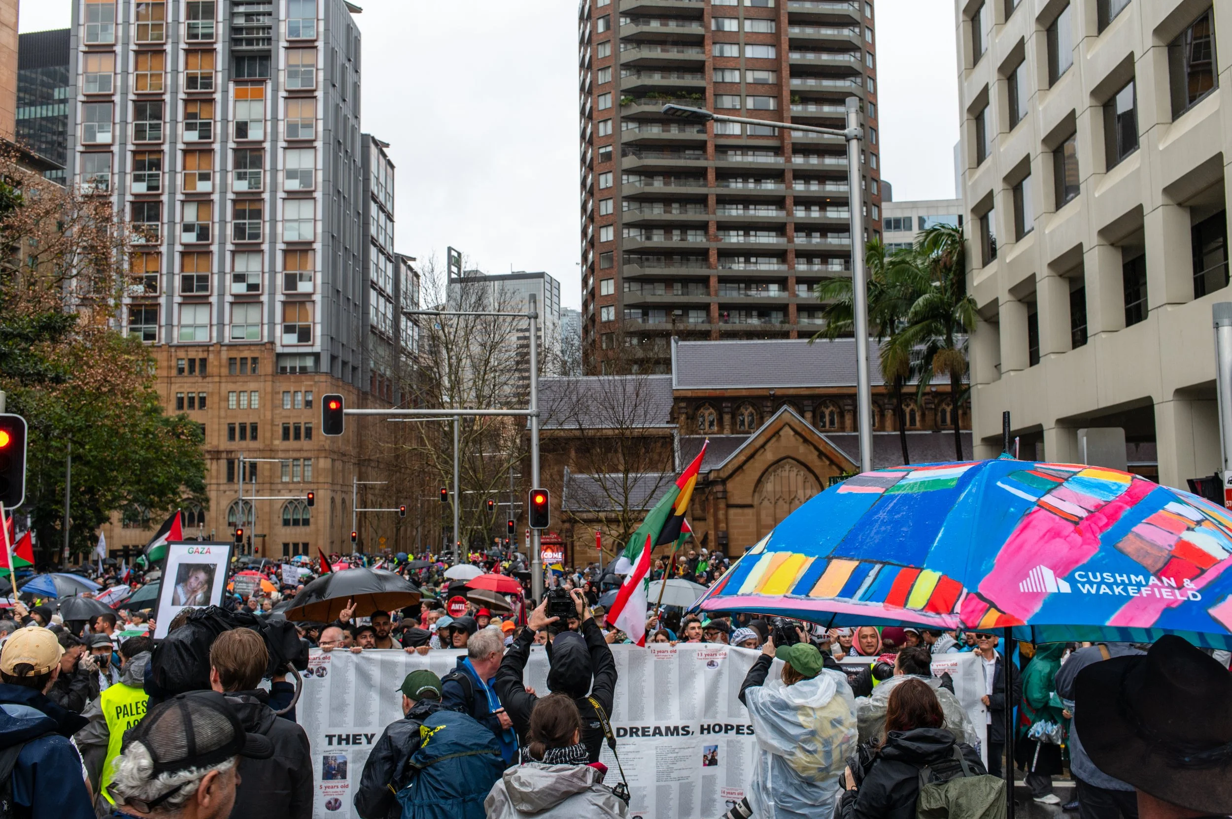 Crowd of protesters holding signs and flags outside in an urban area with tall buildings, some holding umbrellas in rainy weather.