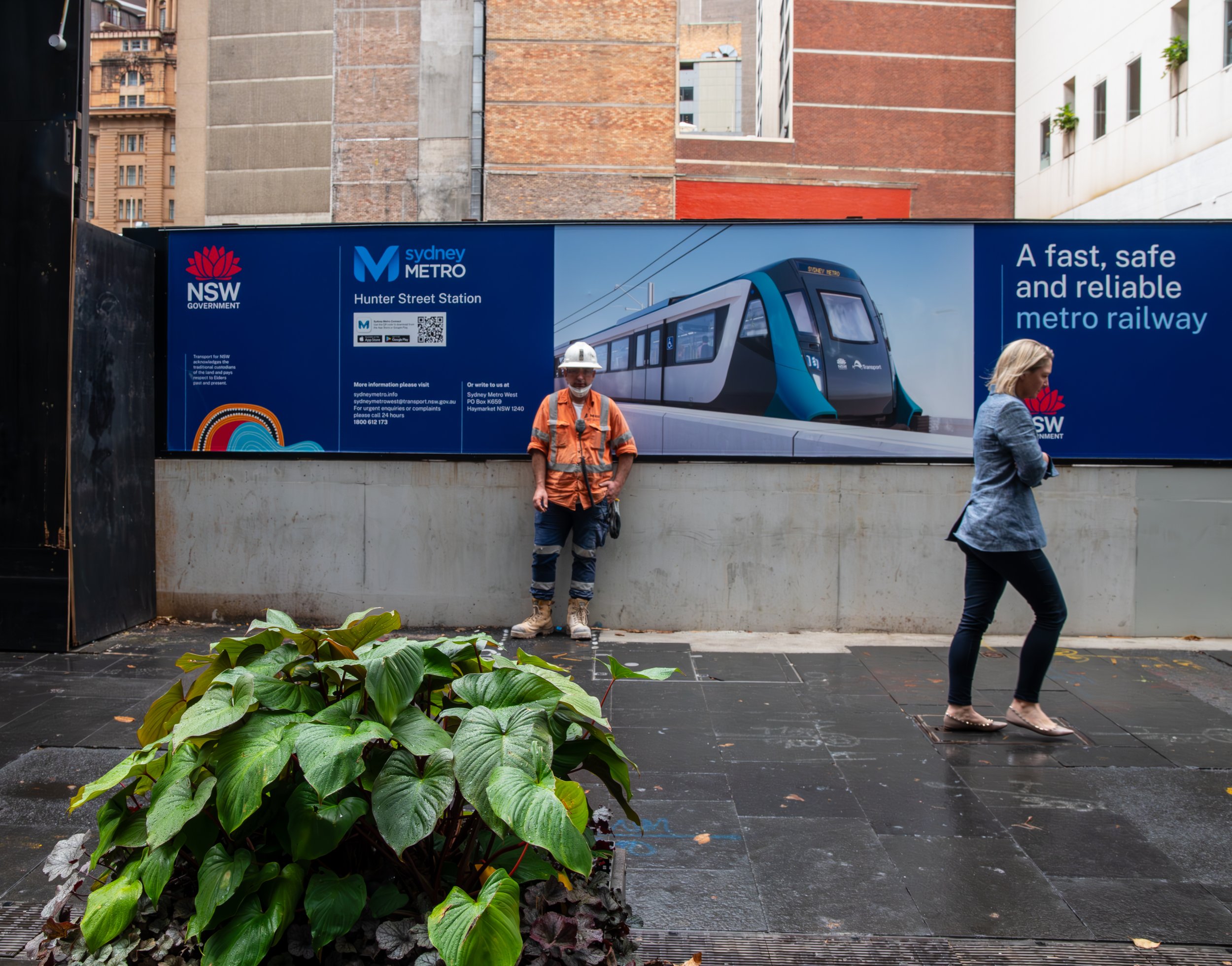 A construction worker in safety gear standing in front of a Sydney Metro advertisement on a city street. A woman is walking past on the sidewalk. The advertisement shows a modern metro train and promotes a fast, safe, and reliable metro railway.