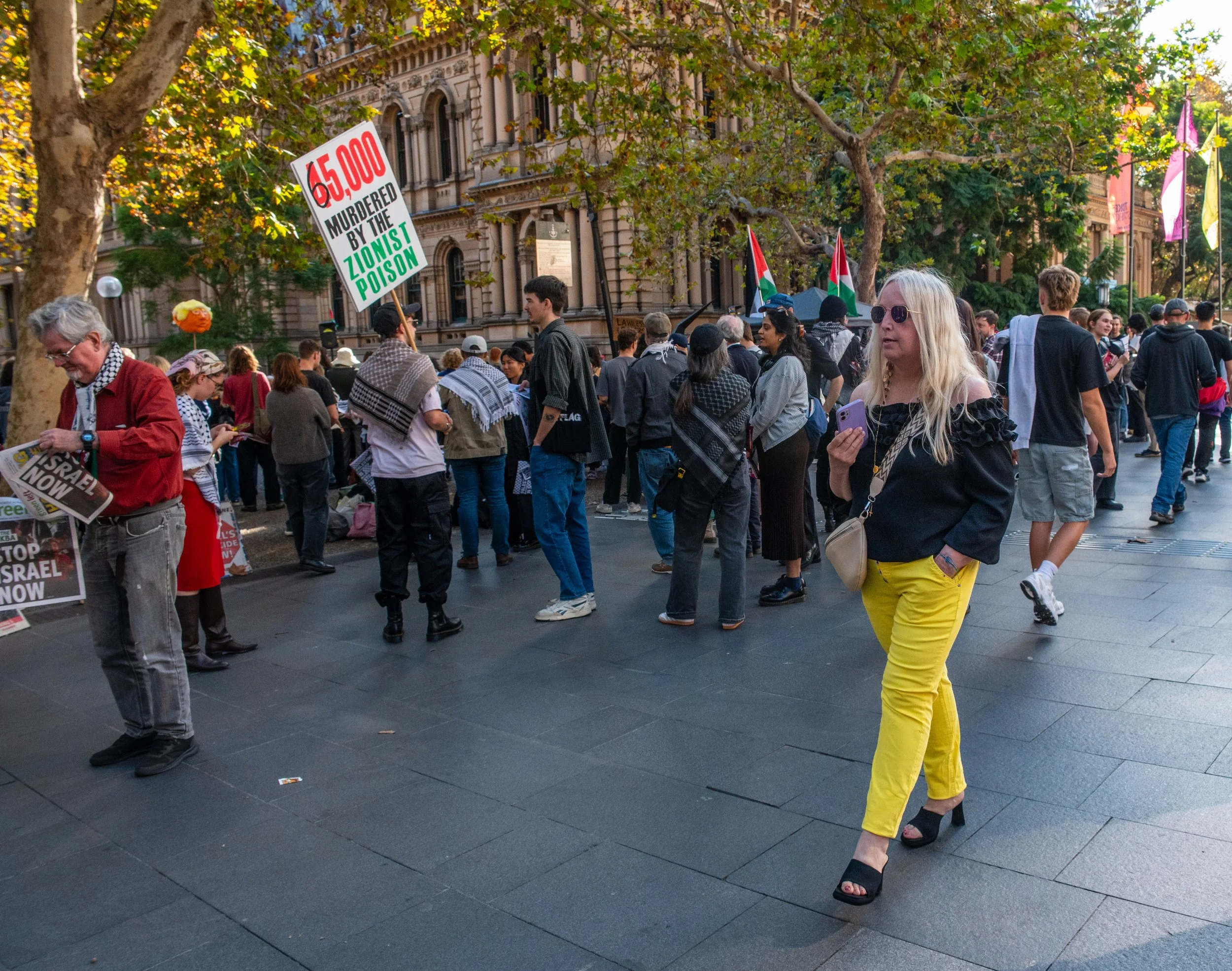 Crowd of protesters holding signs and flags, gathered in front of historic buildings, during a daytime rally.