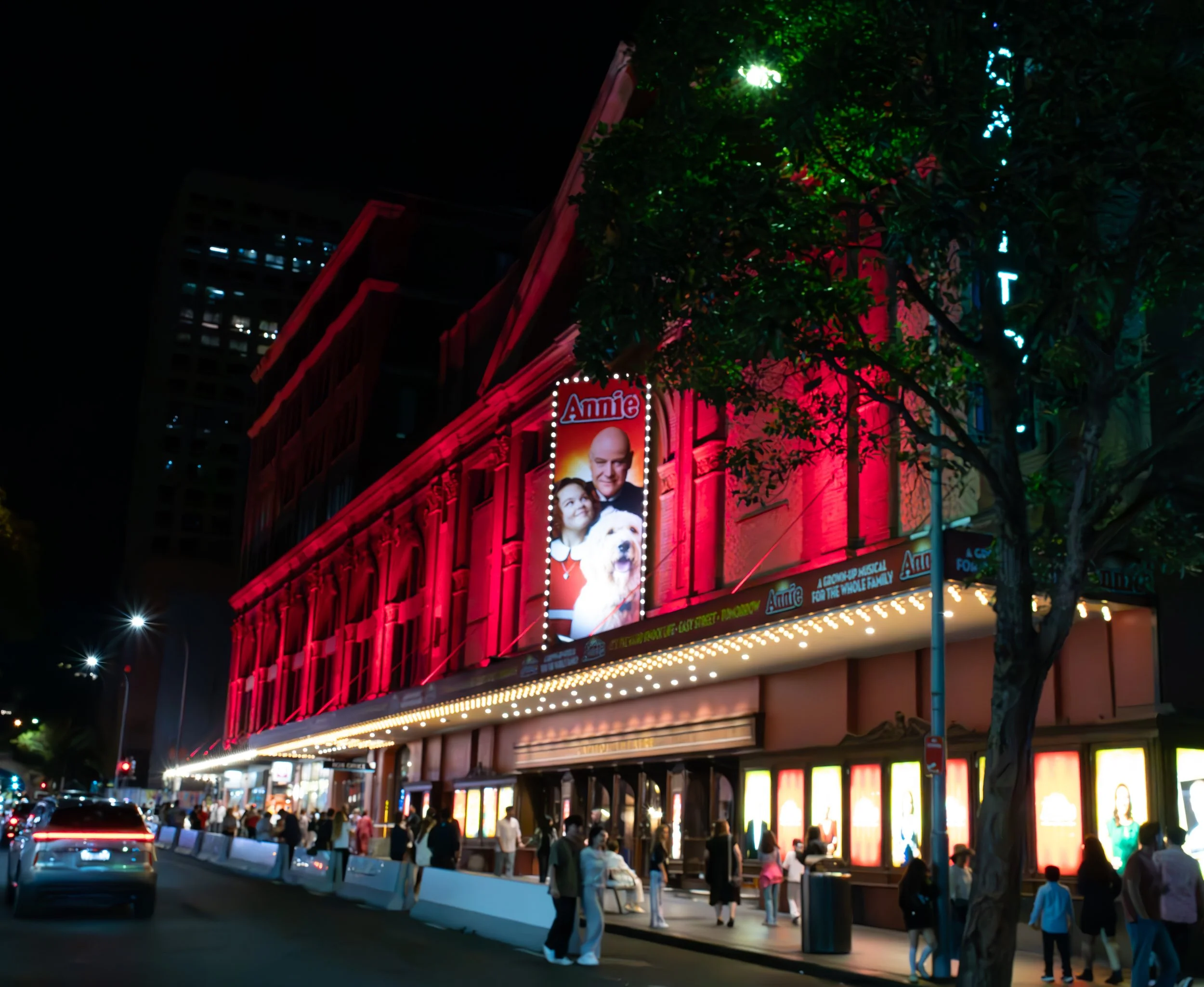 Night view of a theater building decorated with red lighting, Broadway sign, and illuminated posters, with people walking on the sidewalk and cars on the street.