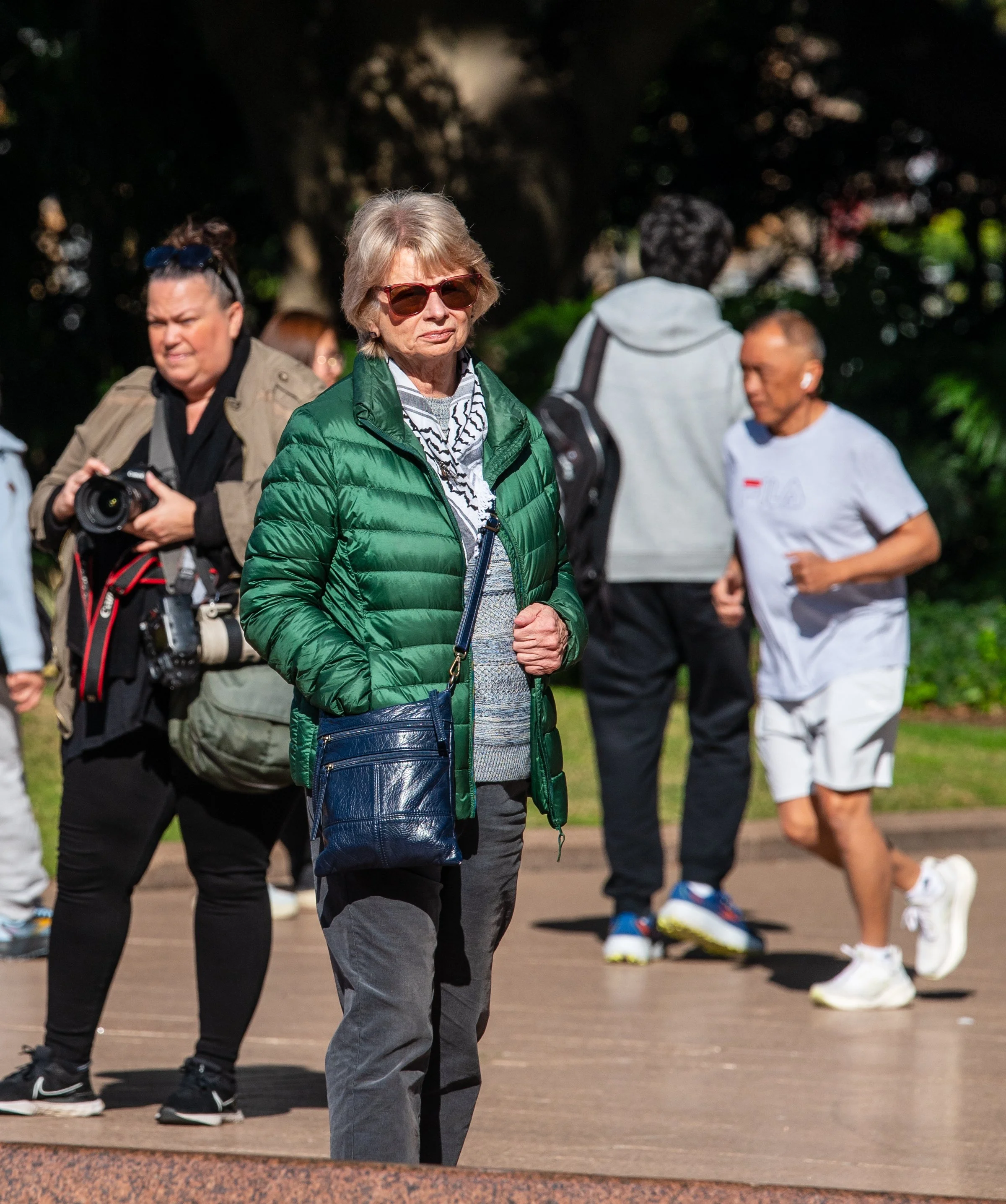 An elderly woman wearing sunglasses and a green puffer jacket stands outdoors among other people in a park or public space during daytime.
