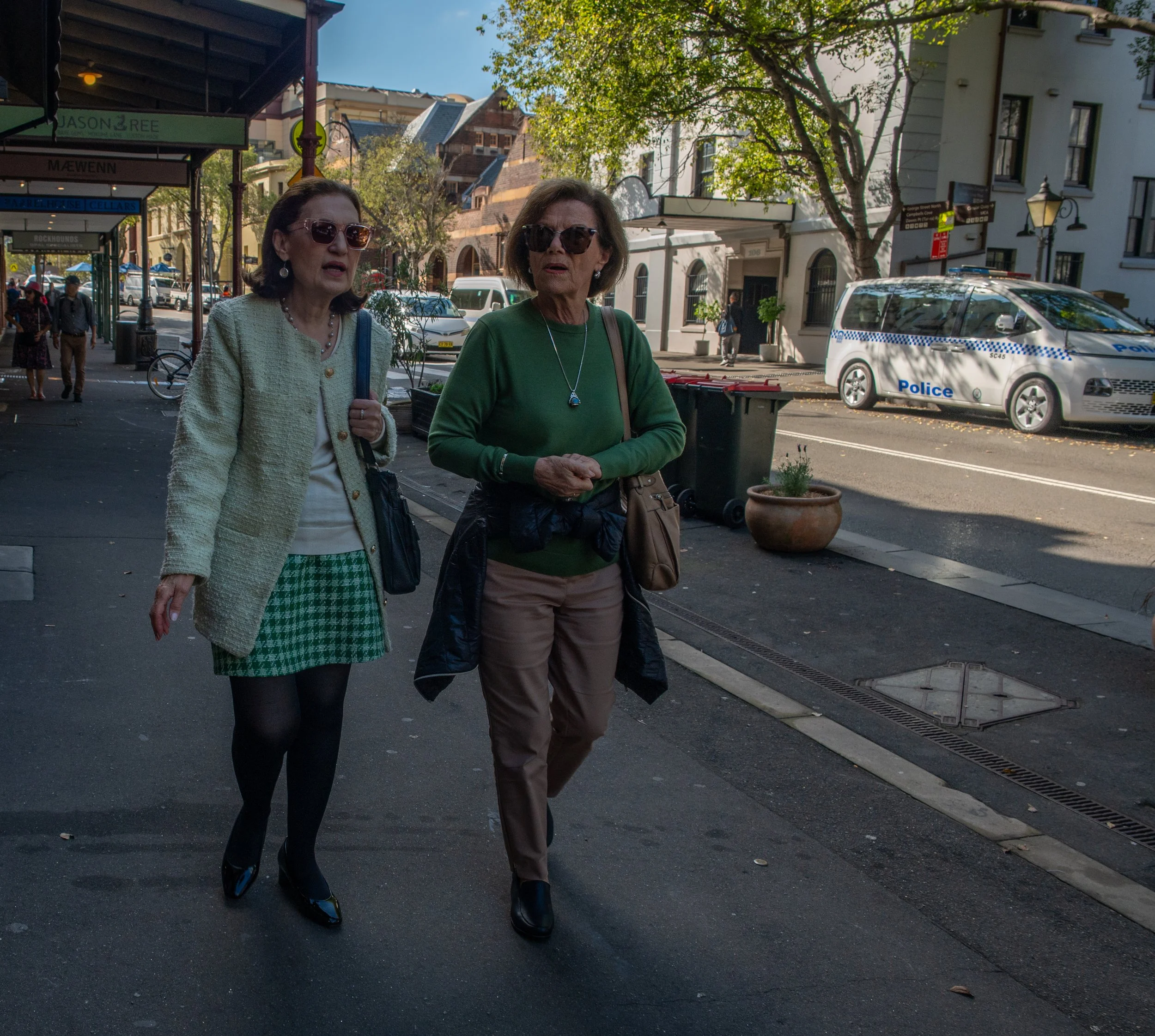 Two women walking on a city sidewalk near parked police car, trees, and storefronts.