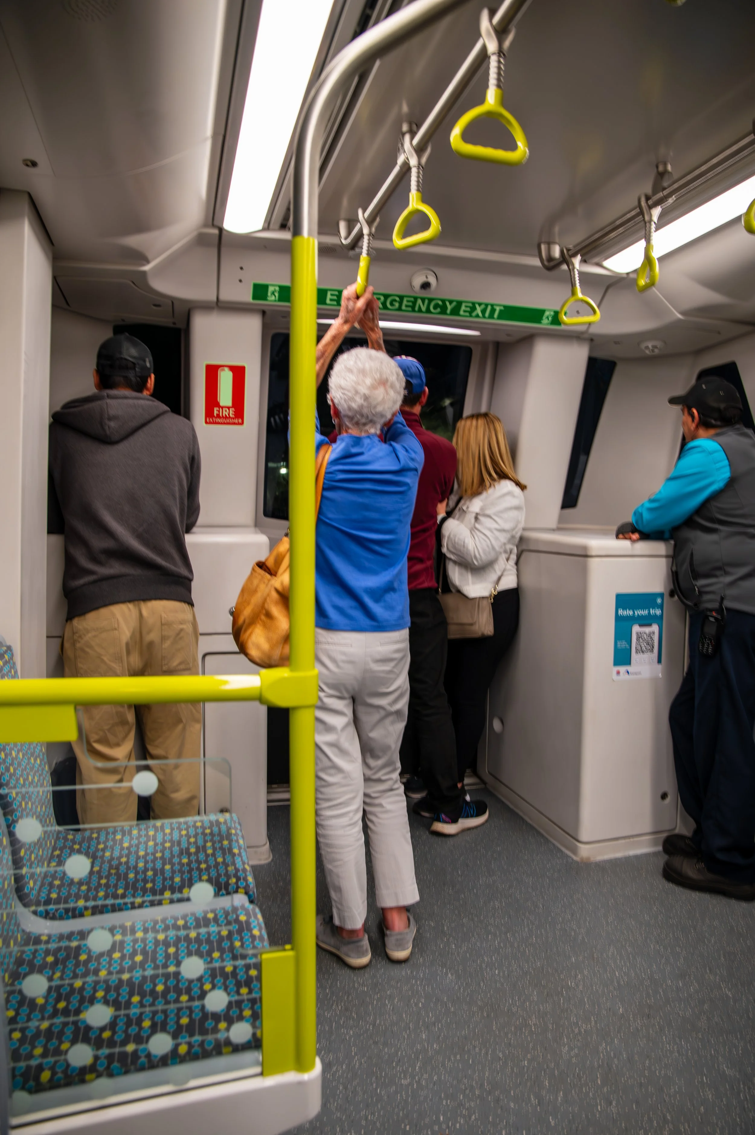 People standing in a line inside a train, with some holding onto handrails and others waiting near the exit door, which has a green emergency exit sign above it.