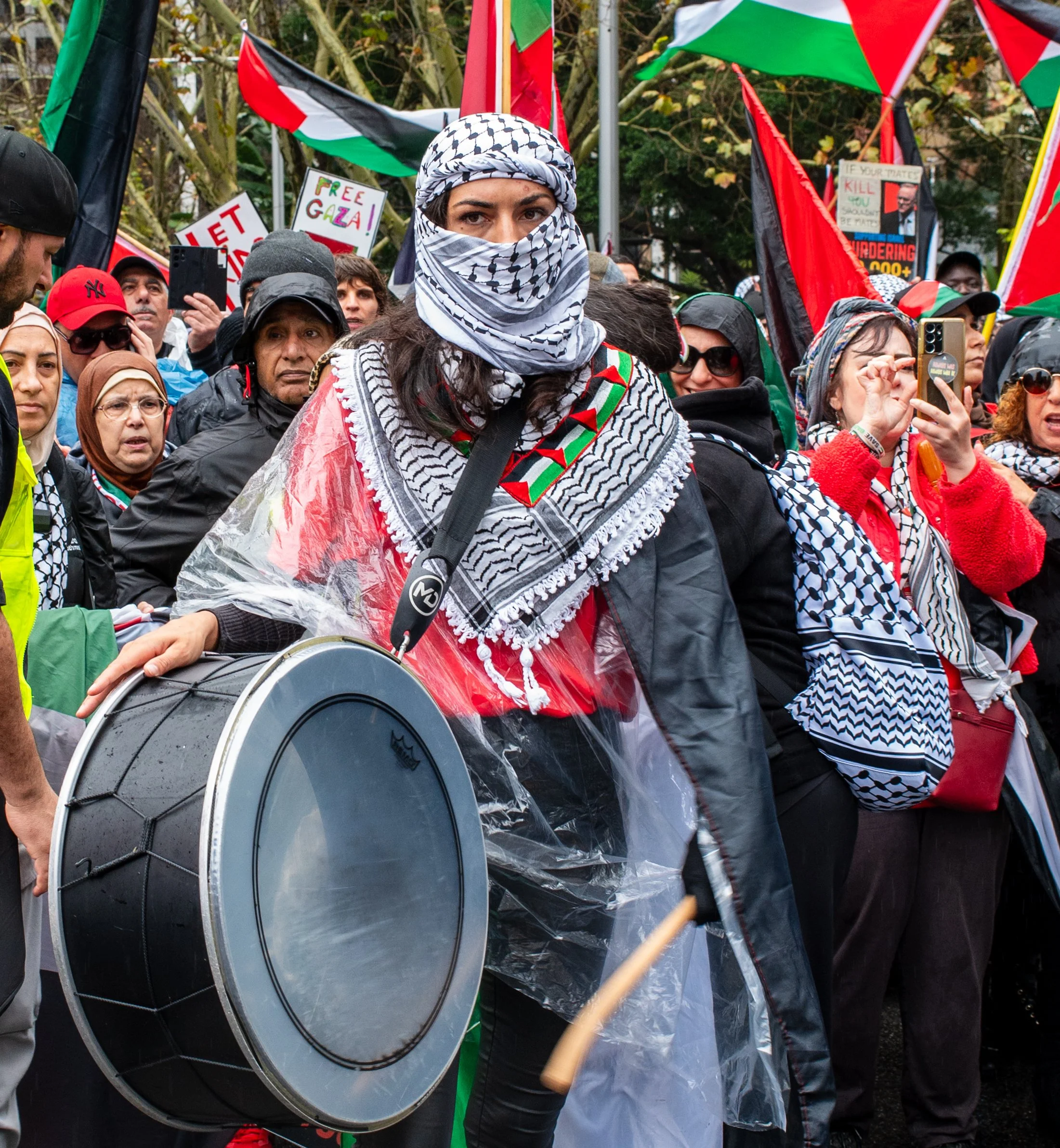 Woman wearing a keffiyeh and a plastic raincoat, holding a large drum at a protest with Palestinian flags and signs in the background.