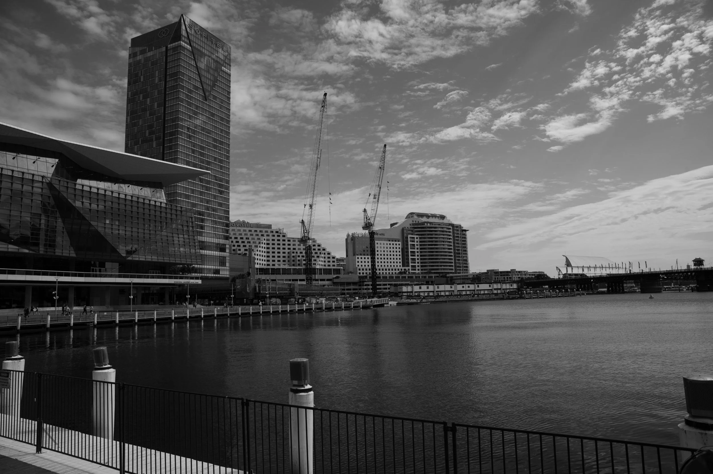 Black and white photograph of a city skyline along a waterfront, featuring modern high-rise buildings, construction cranes, and a bridge over the water, with a sky filled with clouds.