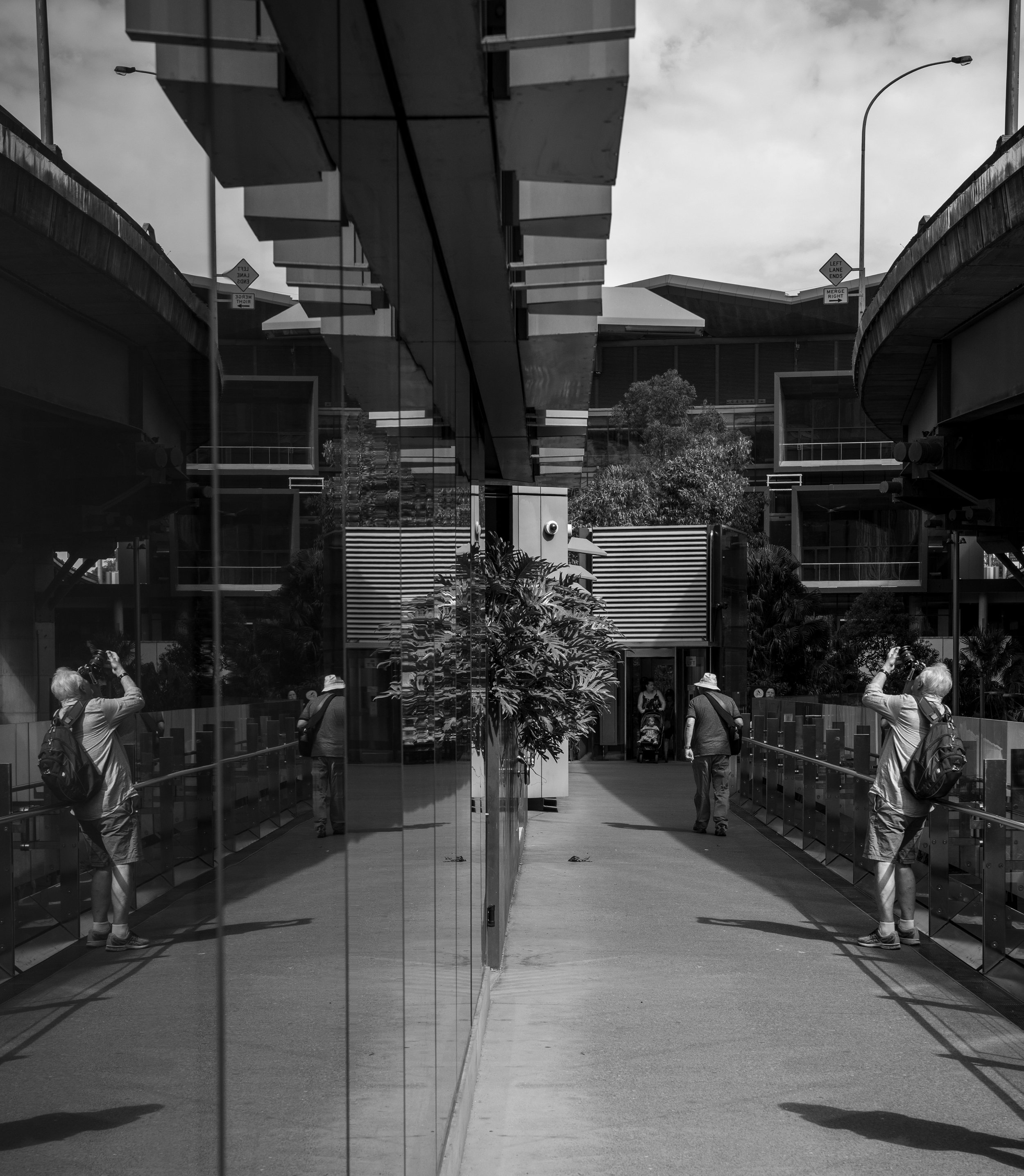 Black and white photo of a modern building with glass walls, featuring reflections of people taking photos and walking. The scene includes trees and a cloudy sky.