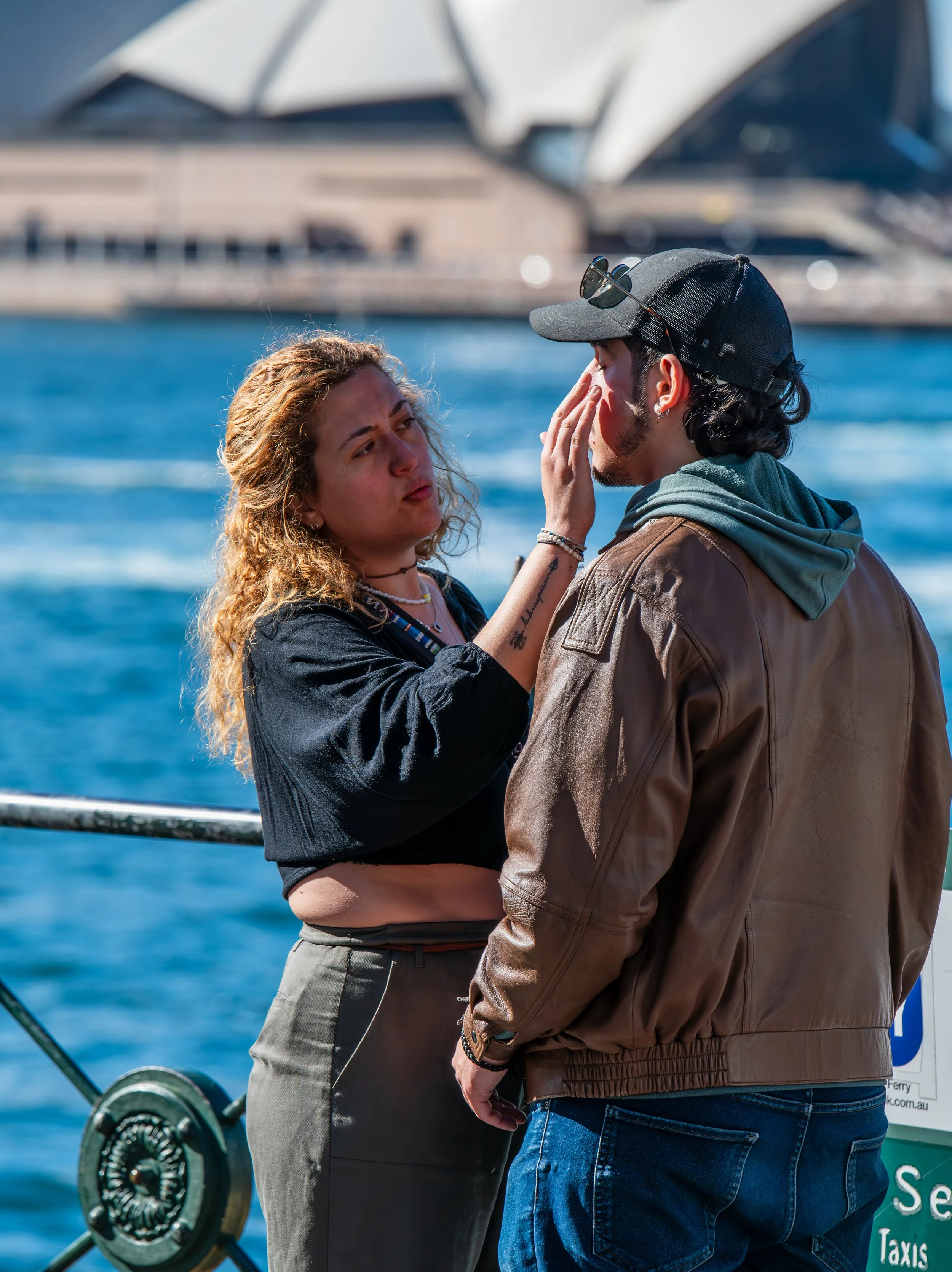 A woman with curly hair and a black top is touching the face of a man wearing a brown leather jacket, green hoodie, black cap, and sunglasses, while standing near the water with the Sydney Opera House in the background.