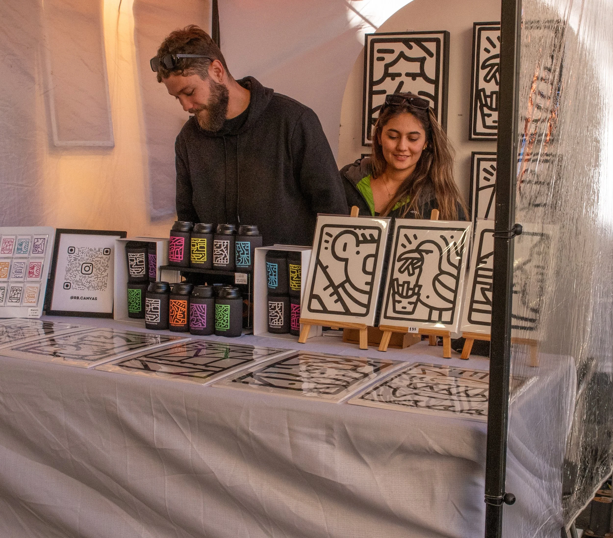 Two people behind a table displaying black and white line art prints and cans with colorful labels at an art or craft market.