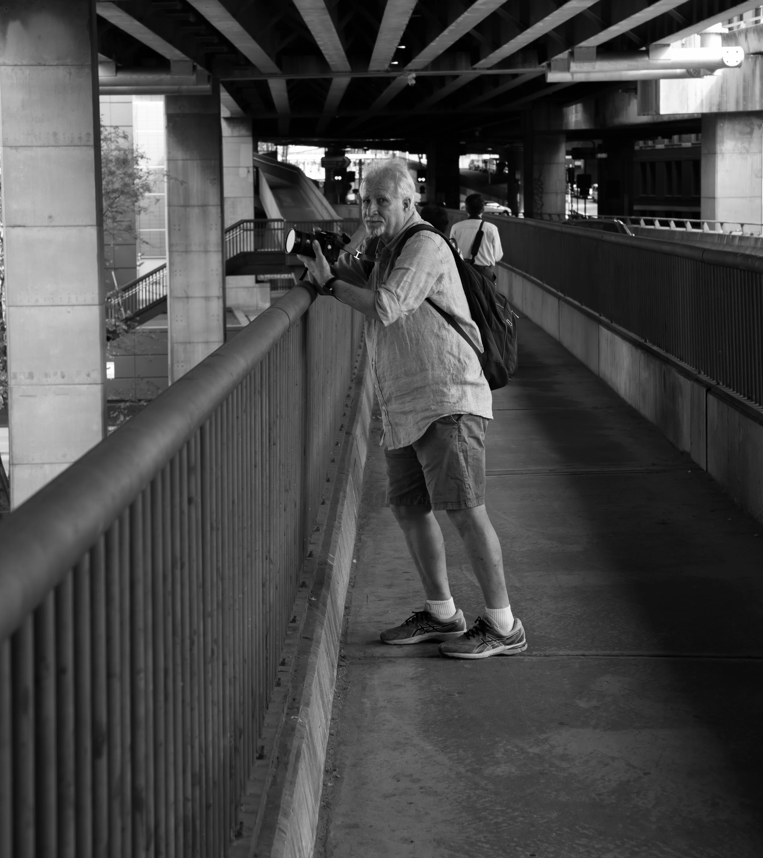 Black and white photo of an older man with a camera, standing on a bridge, looking over the railing. He has a backpack and is dressed in a long-sleeve shirt, shorts, and sneakers. There are other people in the background under an overpass.