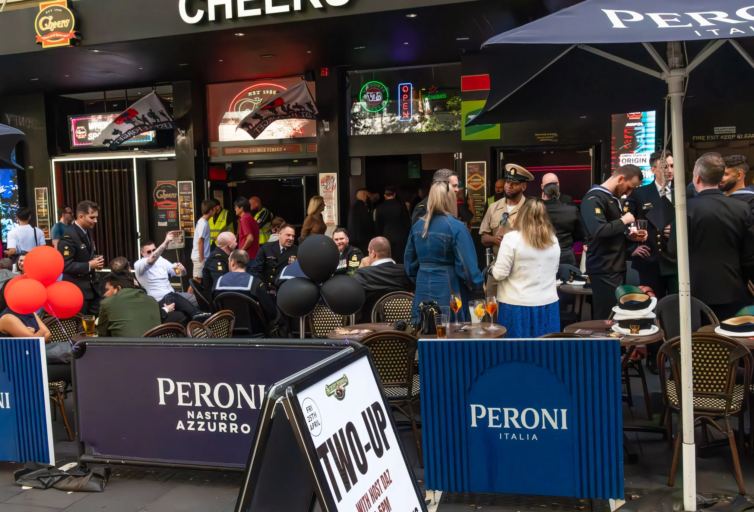People gathered outside a bar or restaurant, some in military uniforms, with decorations including balloons and banners, and a sign that says 'Peroni Italia'.