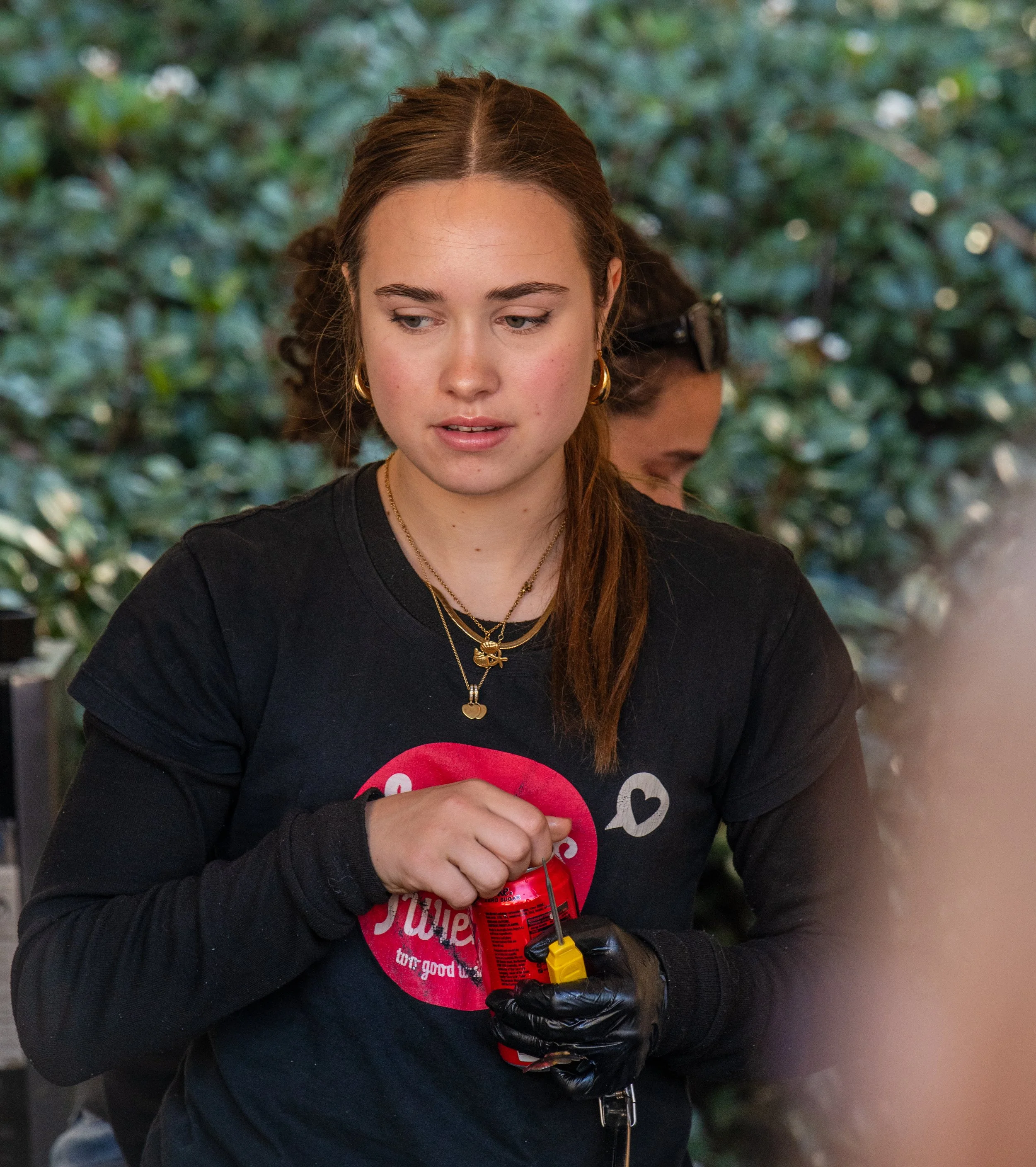 A young woman with brown hair, wearing a black t-shirt with a red graphic, gold jewelry, and a black glove on her left hand, holding a small firecracker with a fuse.