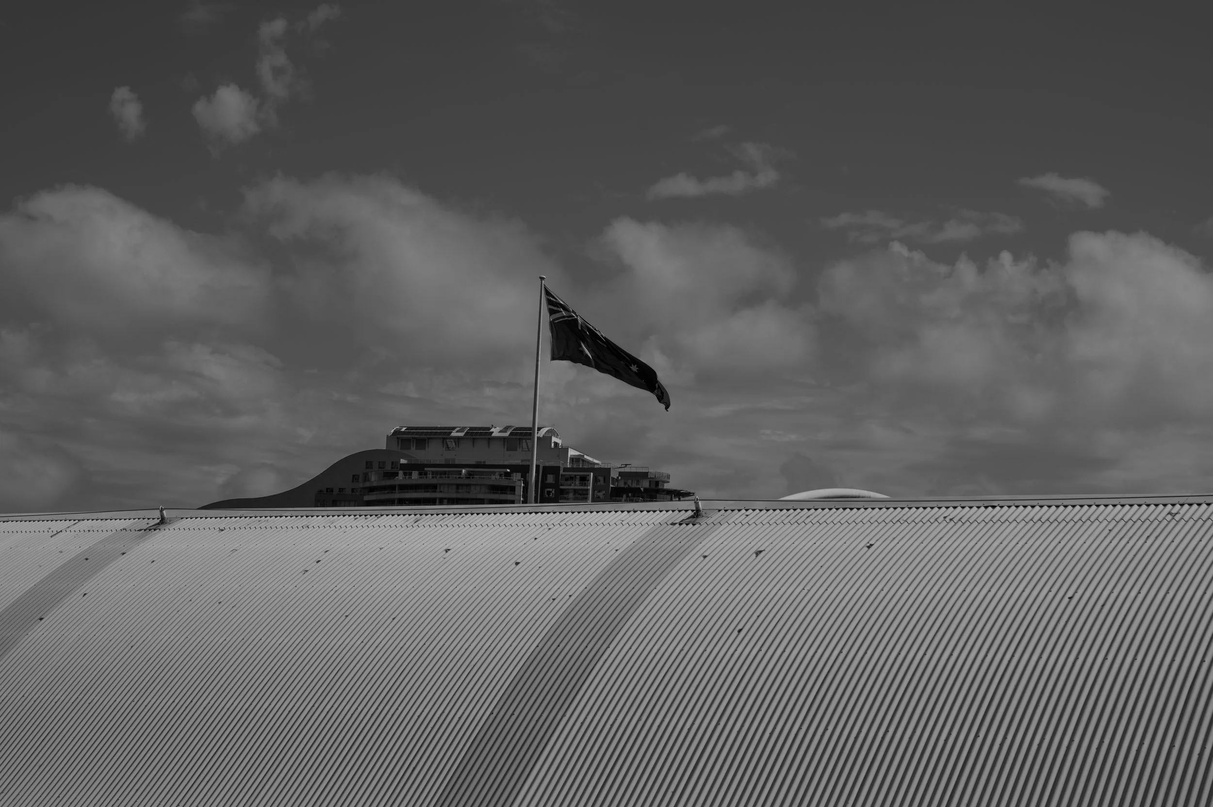 Black and white photo of a flag on a pole atop a corrugated roof, with a modern building and cloudy sky in the background.
