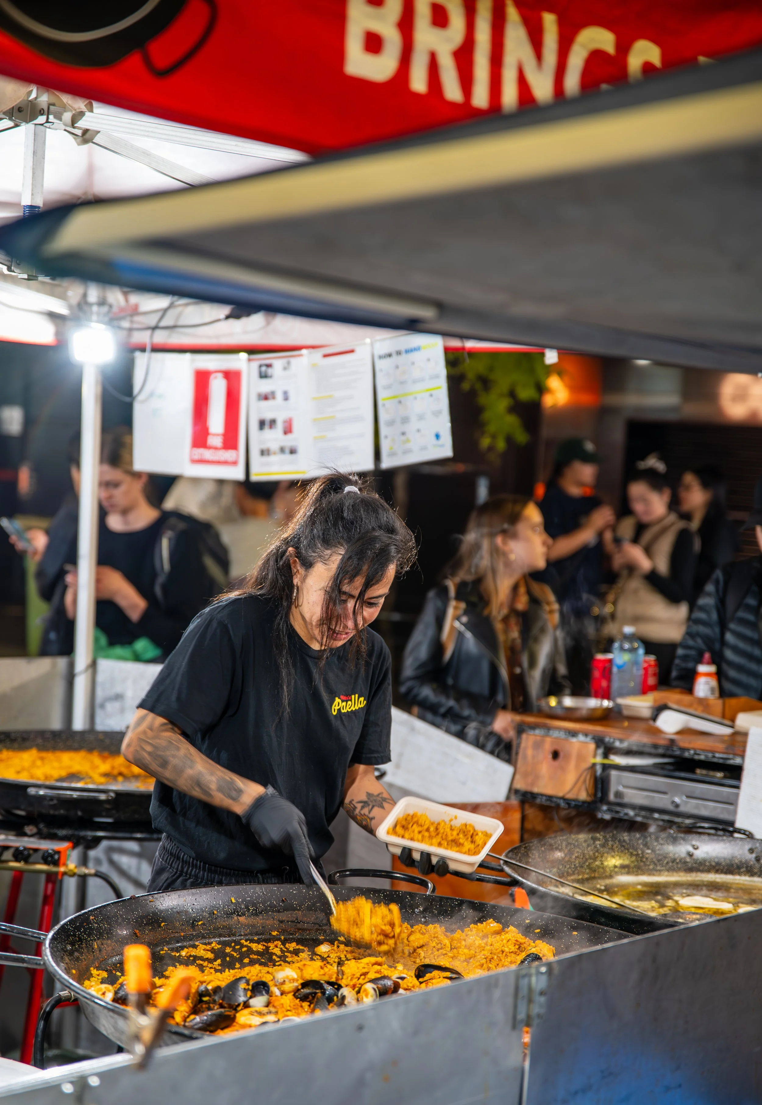 A woman cooking paella at a street food stall, scooping rice from a large pan into a container, with people waiting in line in the background.