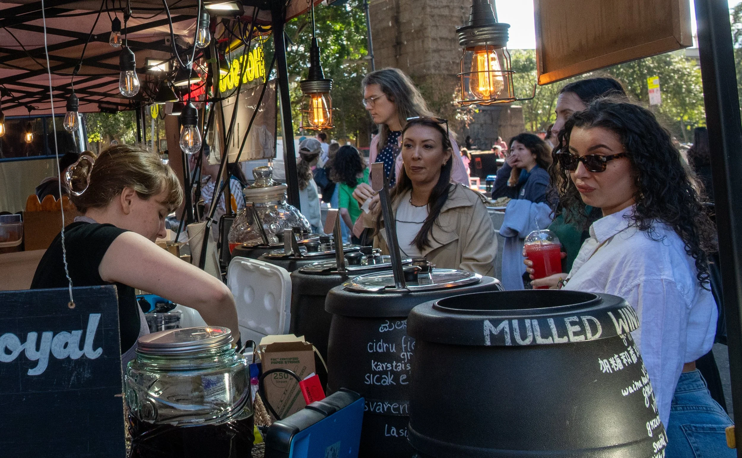 People at an outdoor food stall with hanging light bulbs, ordering food and drinks during daytime.