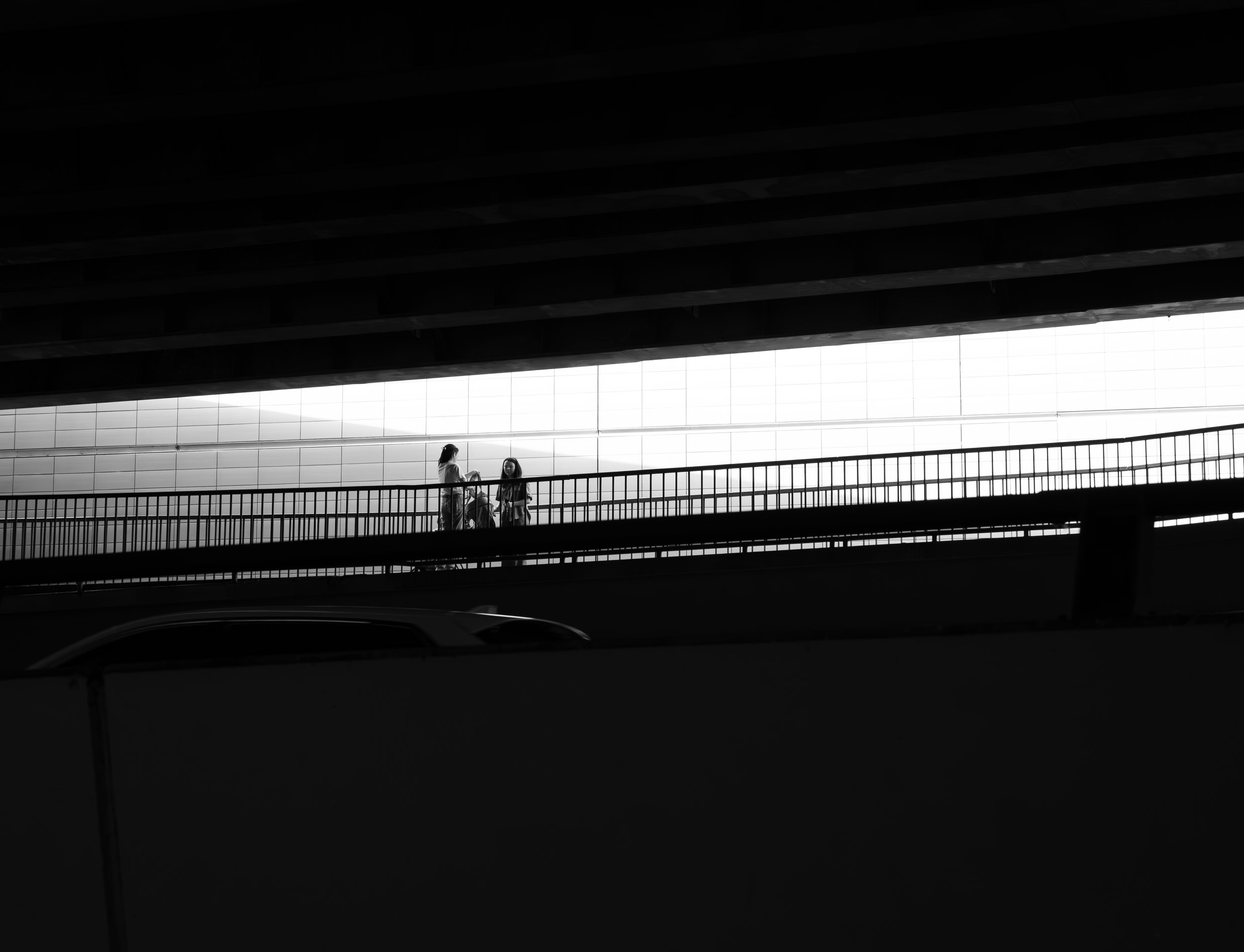 Silhouettes of three people standing on a bridge or elevated walkway underneath a large structure, with a brightly lit grid-patterned window in the background. The scene is in black and white.