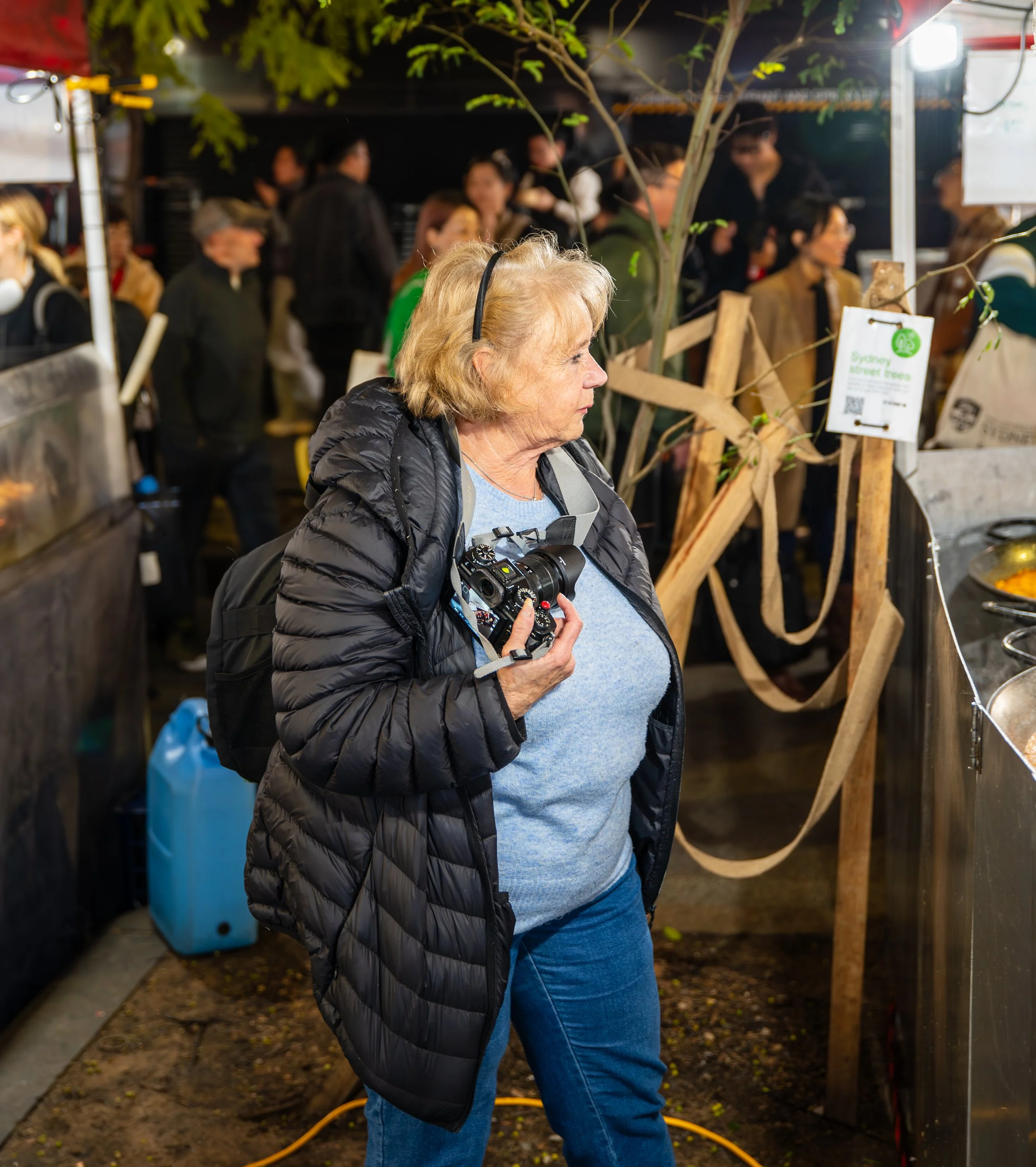 An elderly woman with blonde hair wearing a black puffer jacket, blue shirt, and jeans holding a camera, standing at a busy outdoor market at night surrounded by people and wooden structures.