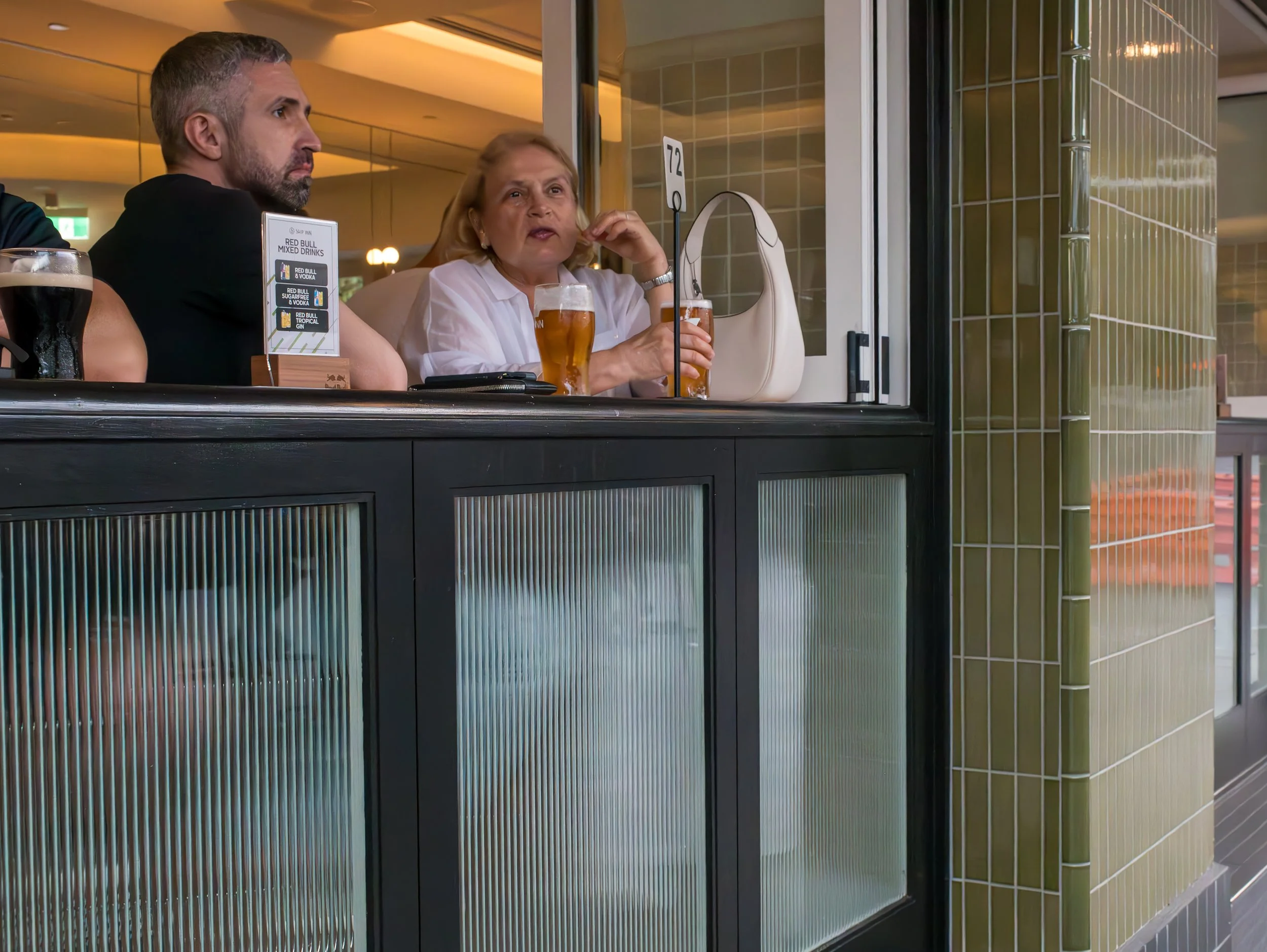 Two people sitting at a bar window, drinking beer and engaging in conversation. The woman is wearing a white shirt, and the man has gray hair and a beard. There is a small sign on the counter with drink options.