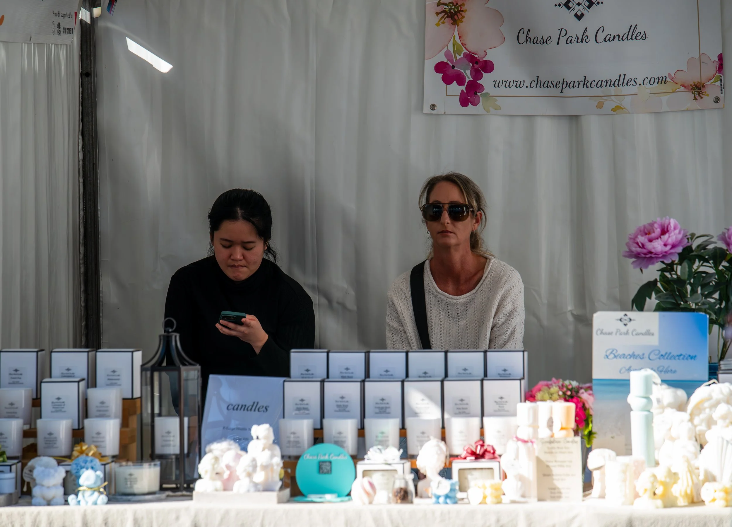 Two women at a booth selling candles, with one looking at her phone and the other wearing sunglasses, behind a table with various candles and a sign that reads 'candles' and 'Chase Park Candles'.