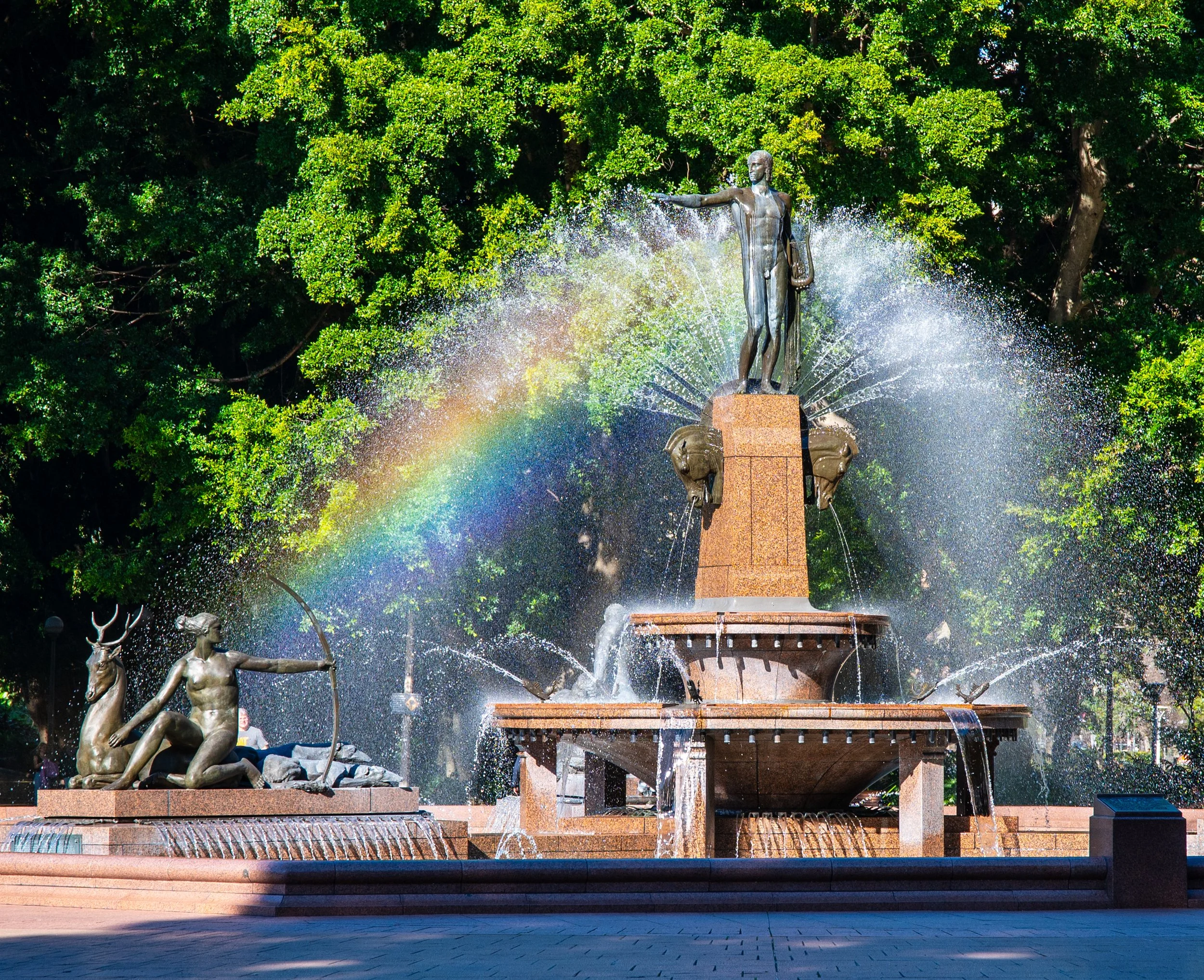 A decorative fountain with sculptures of a woman holding a bow and a reindeer, and a man with a jug, with water spraying and a rainbow in the background, surrounded by lush green trees.