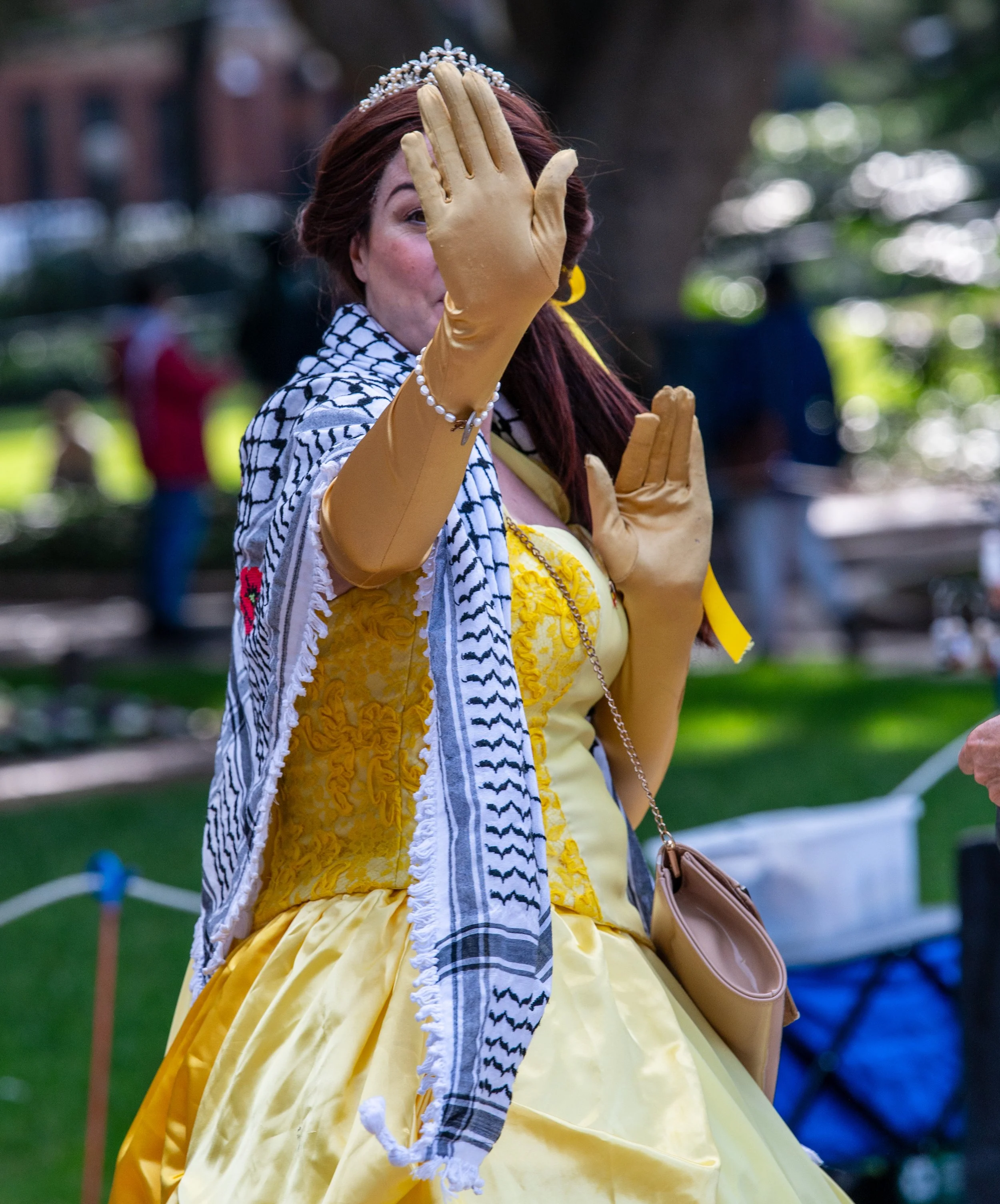 A woman dressed in a yellow gown, wearing a tiara, yellow gloves, and a white and black keffiyeh scarf, making a peace sign with her hand while outdoors during daytime.