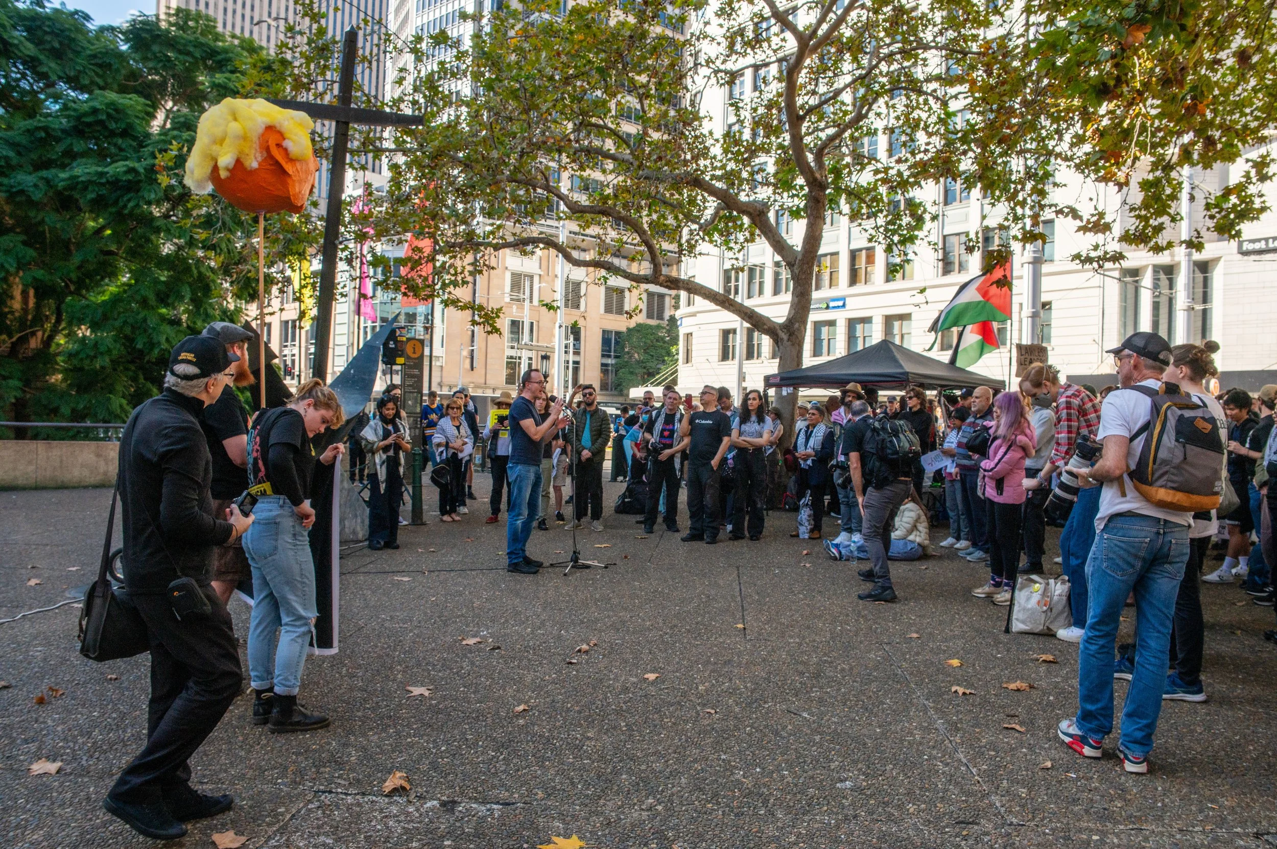 Group of people gathered outdoors in an urban park, some are standing and listening to a person speaking into a microphone, with a tent and flags, including a Palestinian flag, in the background. There is a large tree overhead and a floating paper mache head with blonde hair to the left.