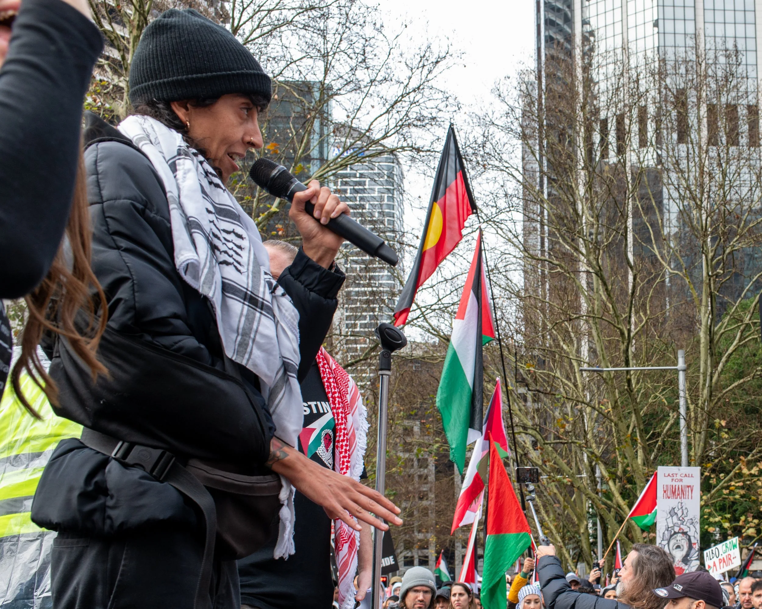 A woman speaking into a microphone at an outdoor protest, with flags and a crowd in the background.