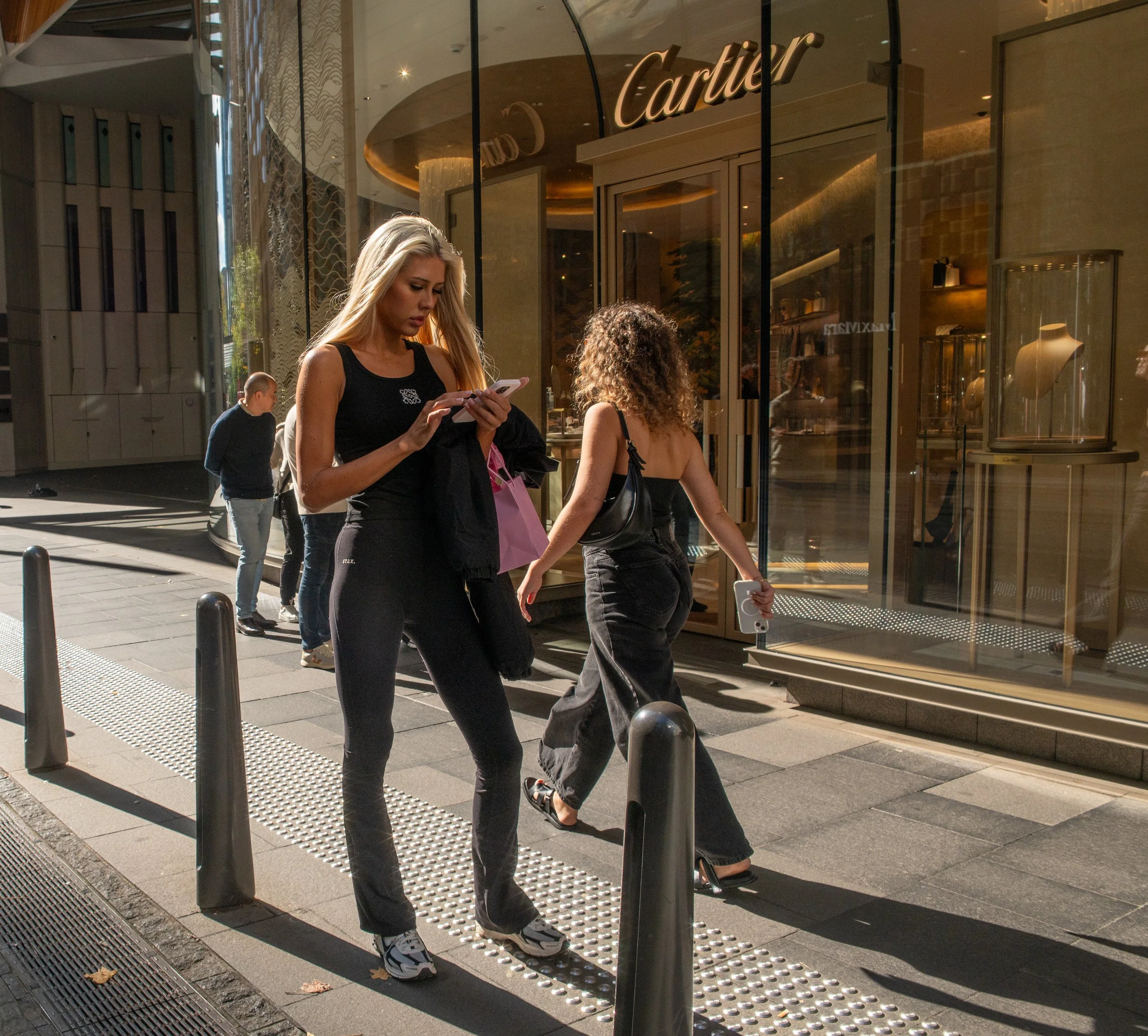 Two women walking outside a Cartier store, one looking at her smartphone, and others walking in the background.