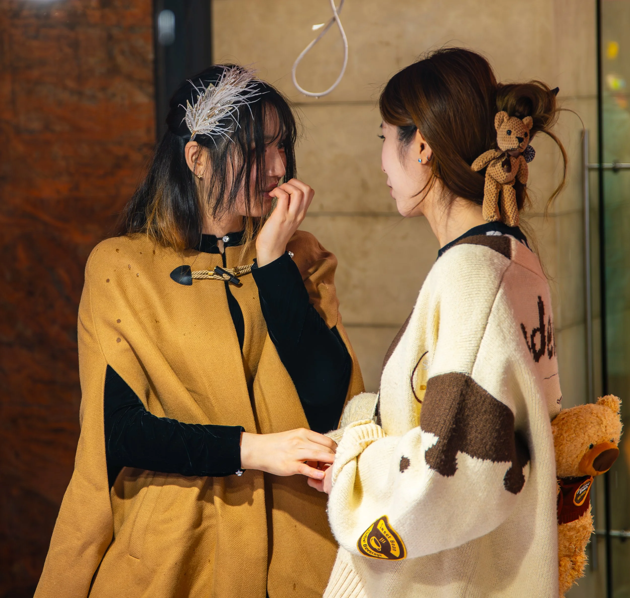 Two women standing close and engaging in a conversation indoors. One woman has shoulder-length black hair with a white feathered hair accessory, wearing a tan coat with black sleeves. The other woman has brown hair styled with a teddy bear hair clip and is wearing a cream and brown sweater with a teddy bear backpack.