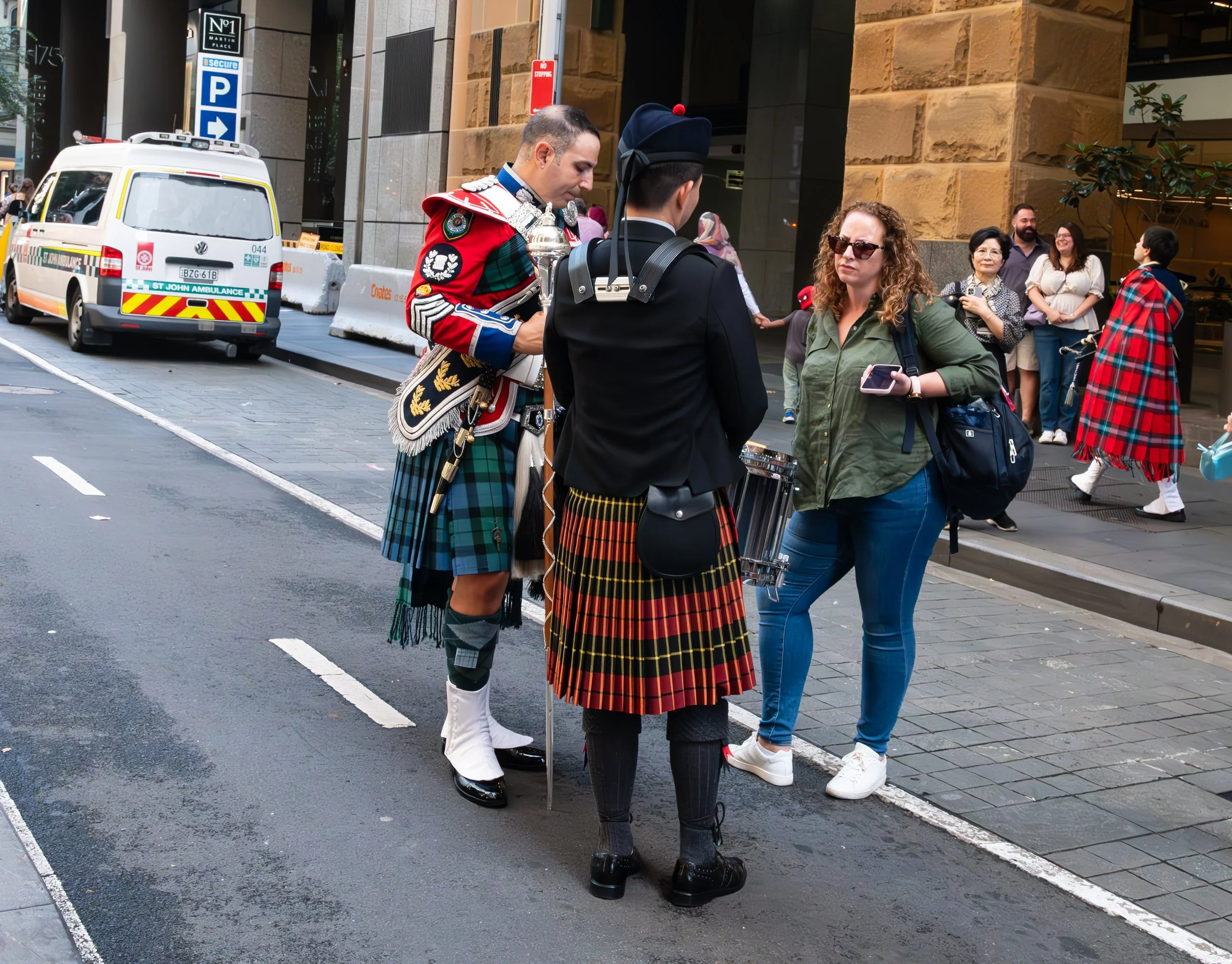 A woman standing on a city sidewalk, talking to two men dressed in traditional Scottish Highland dress, with a group of pedestrians in the background and an ambulance parked nearby.