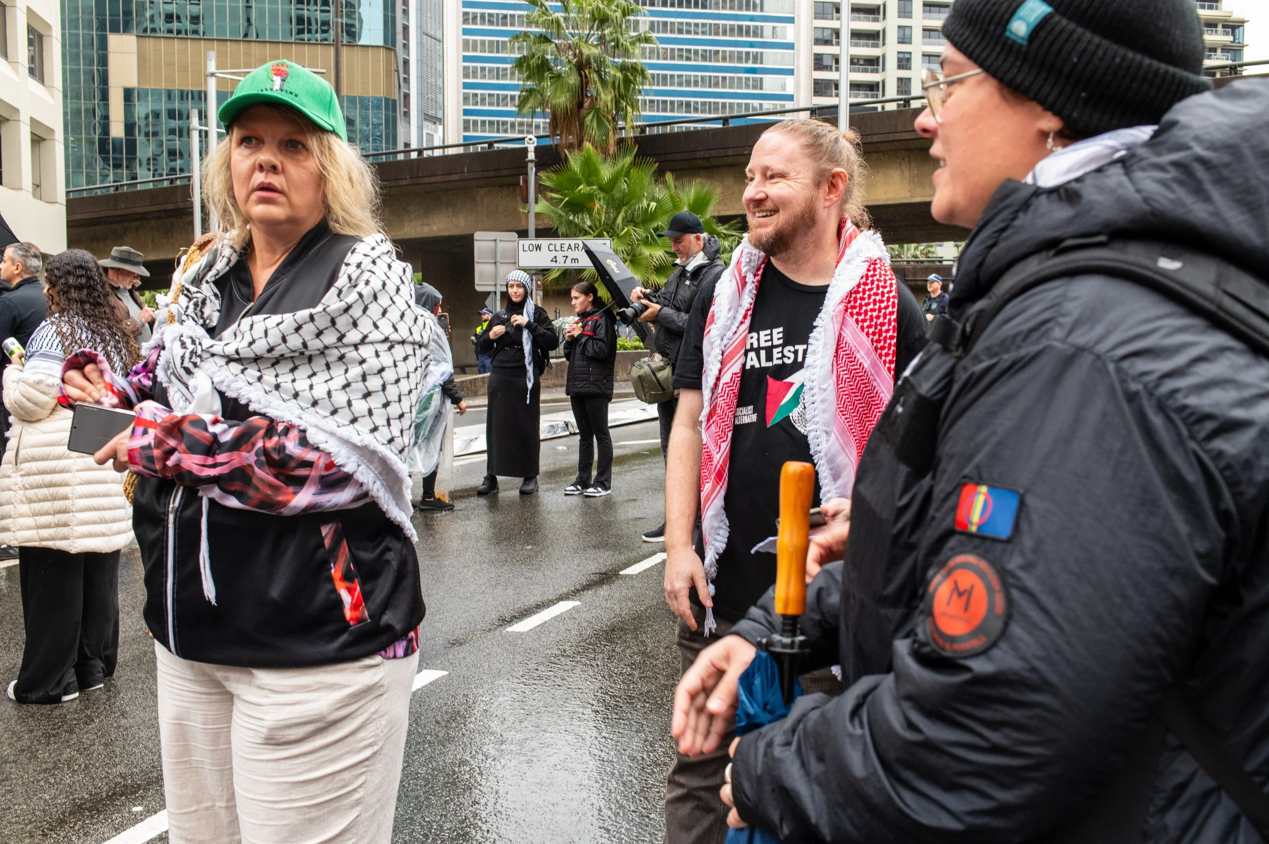 A group of protesters on a city street during a demonstration. One woman in the foreground wears a green cap and a keffiyeh scarf, holding a mobile phone, while another woman in the background is smiling and wearing a red and white keffiyeh scarf with a shirt promoting free Palestine. Other individuals are visible in the background, some with cameras, amidst skyscrapers and palm trees, with wet pavement indicating recent rain.