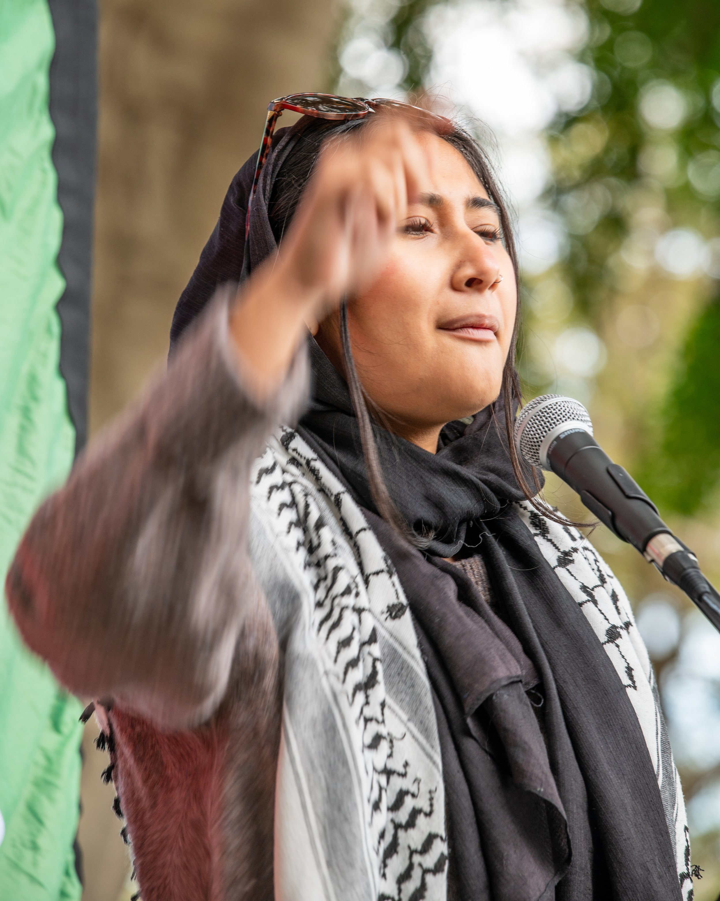 A woman with long dark hair, wearing a black head covering, scarf, and patterned shawl, speaking into a microphone outdoors with trees in the background.